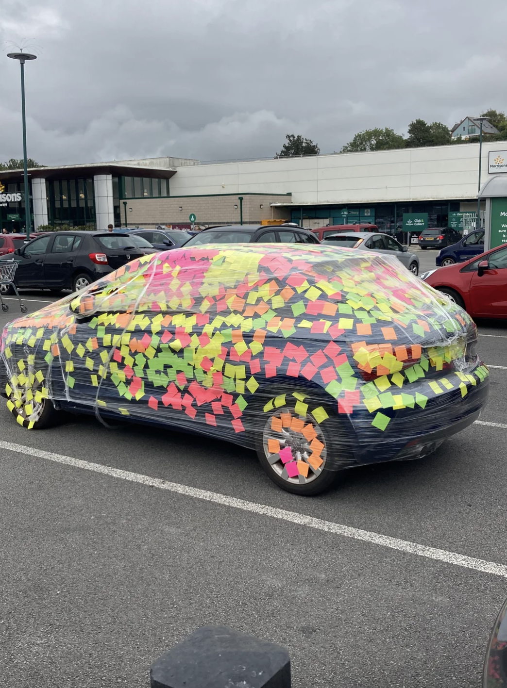 Car covered in multicolored sticky notes and plastic wrap in a parking lot