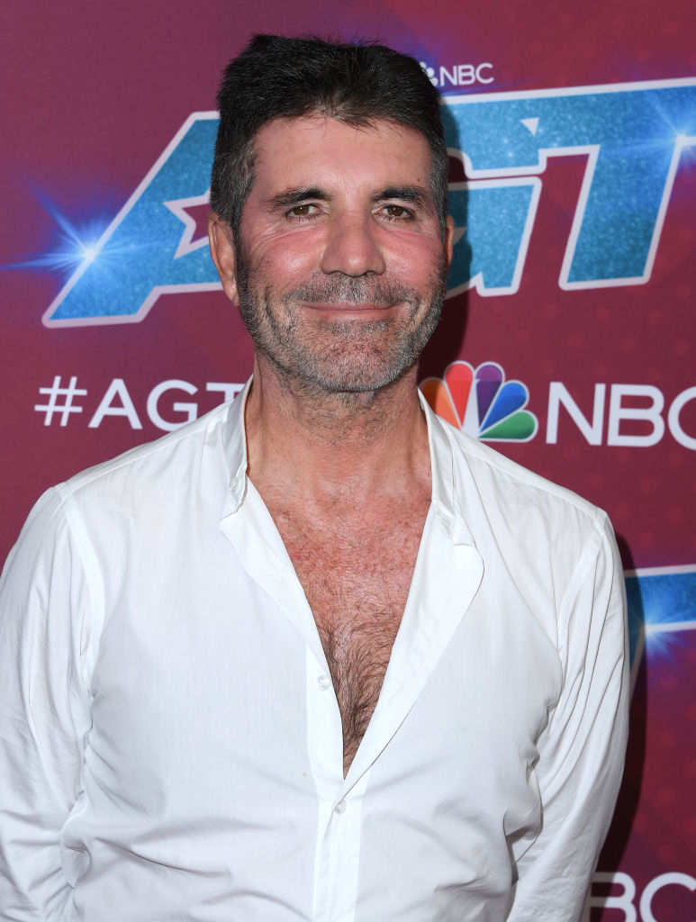 Simon standing in front of an &quot;America&#x27;s Got Talent&quot; backdrop, wearing an open white shirt