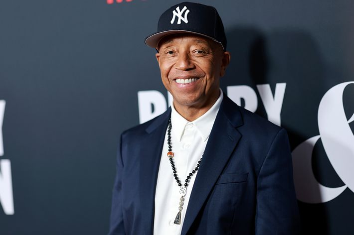 Russell Simmons wearing a blazer and baseball cap with a beaded necklace, smiling at an event