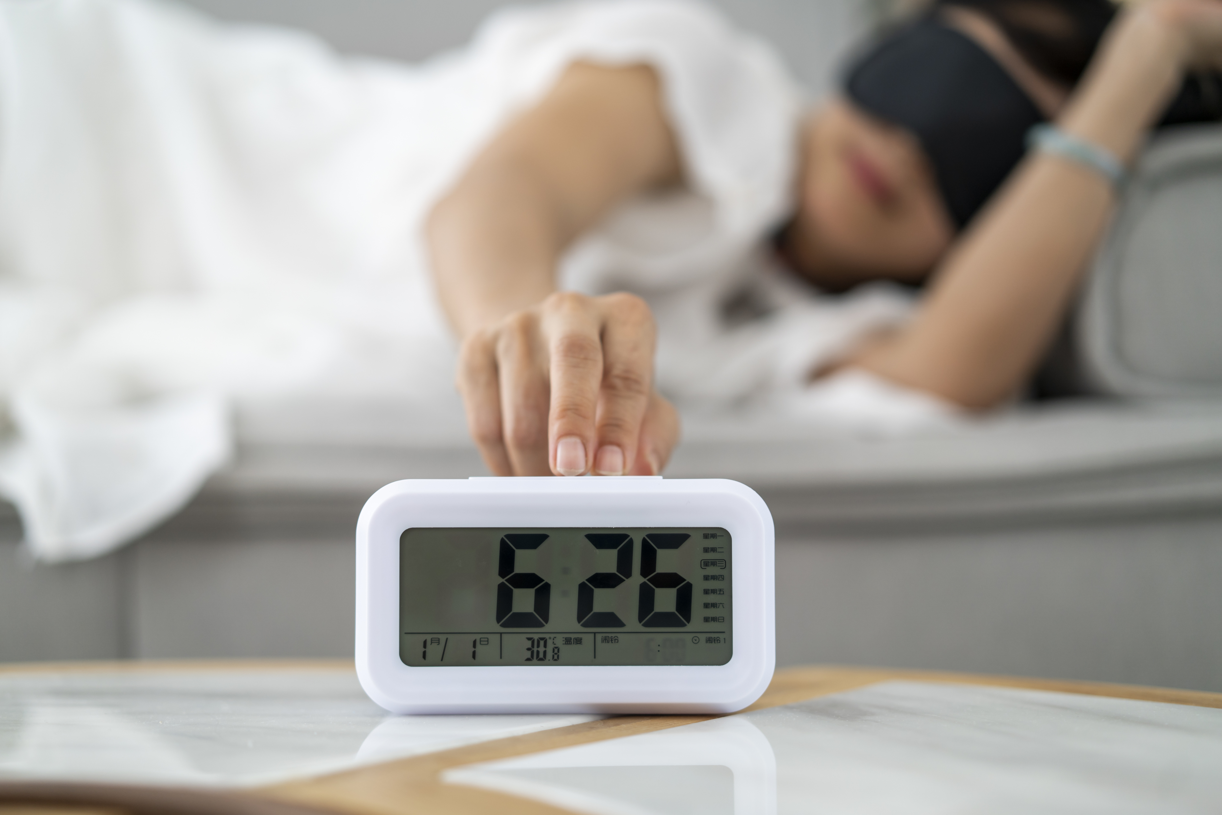 Person in bed reaching out to silence digital alarm clock reading 6:28