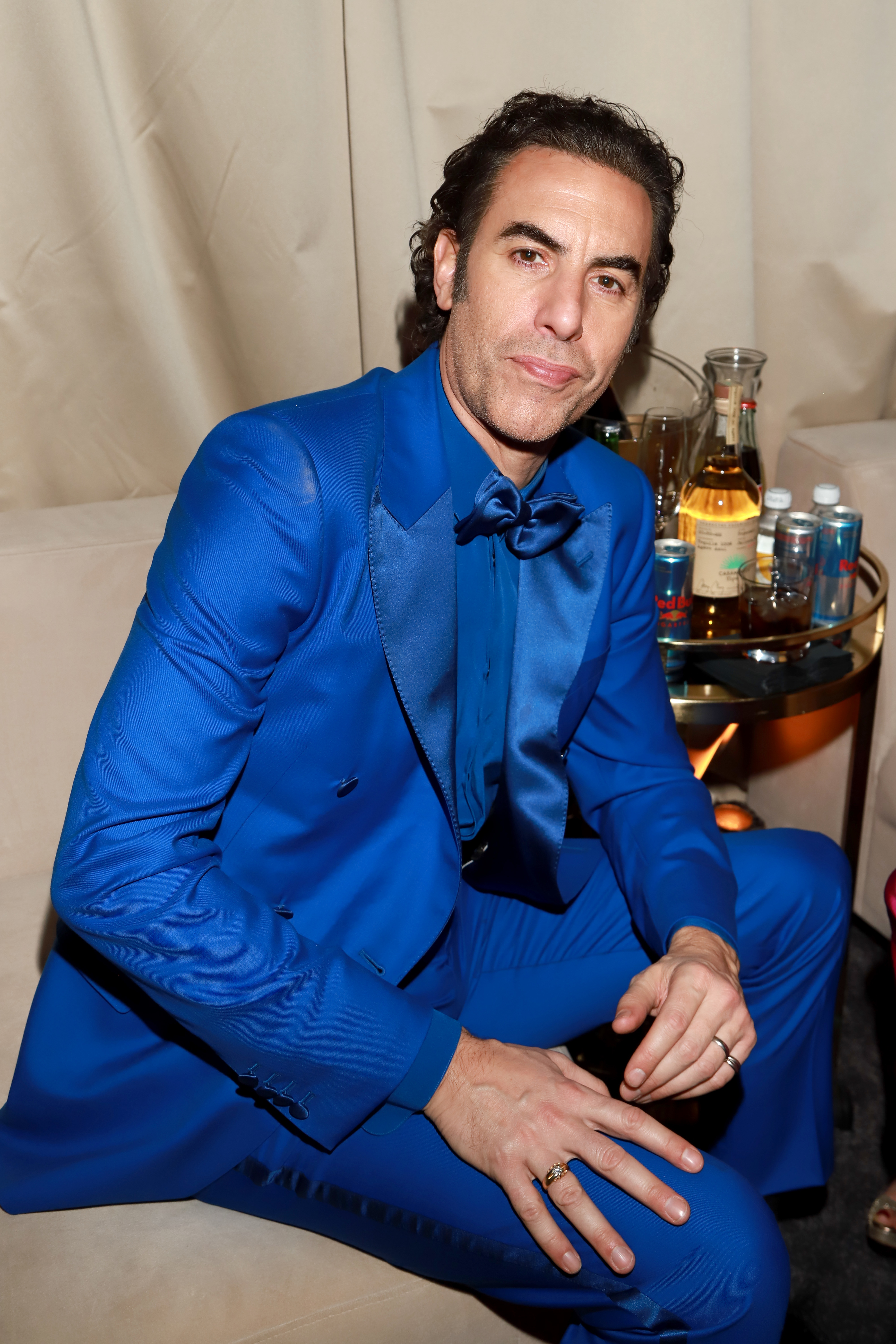 Sacha Baron Cohen in a suit seated with a bow tie at an event