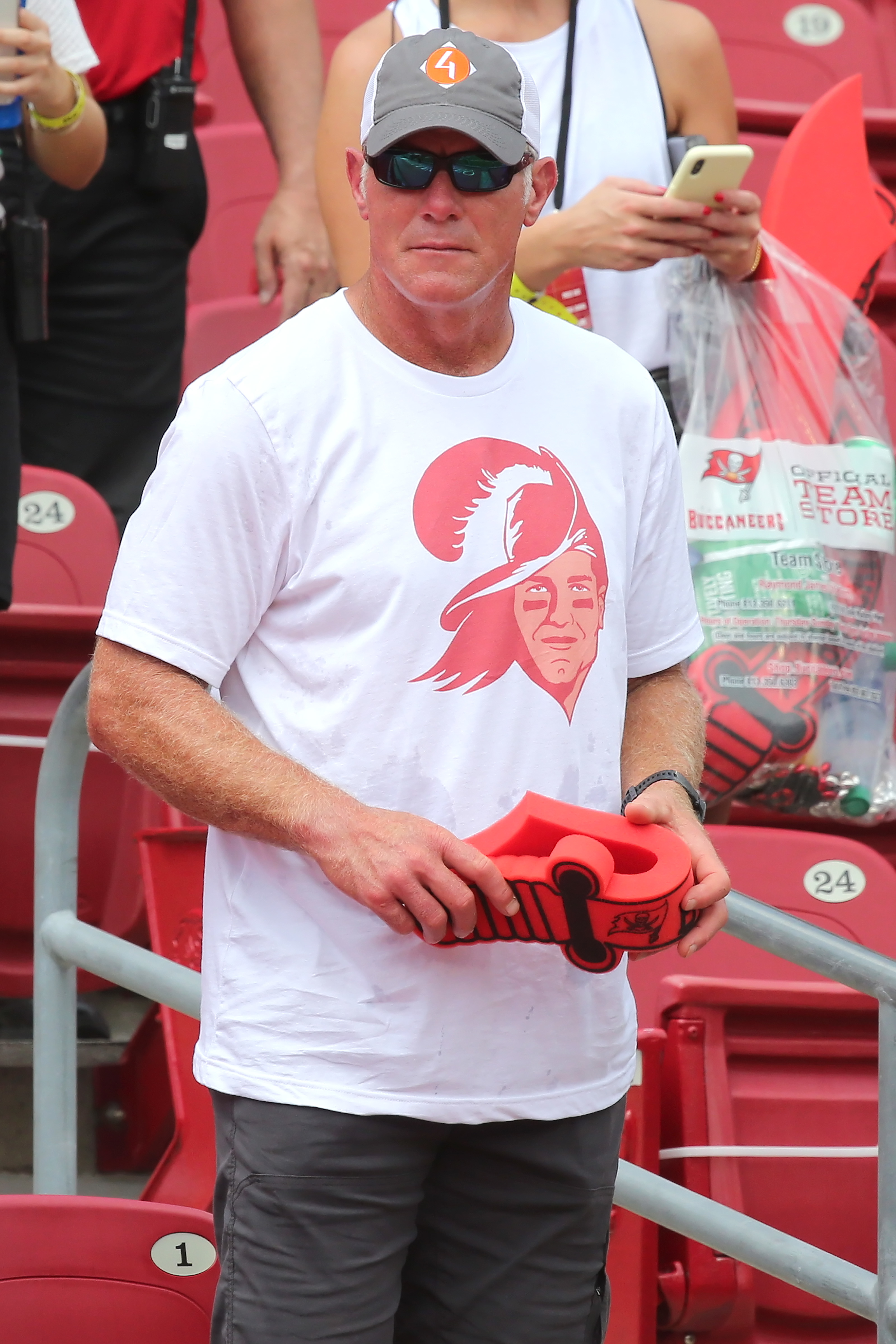 Brett in a  cap and white T-shirt with graphic, holding a red object, standing at a sporting event