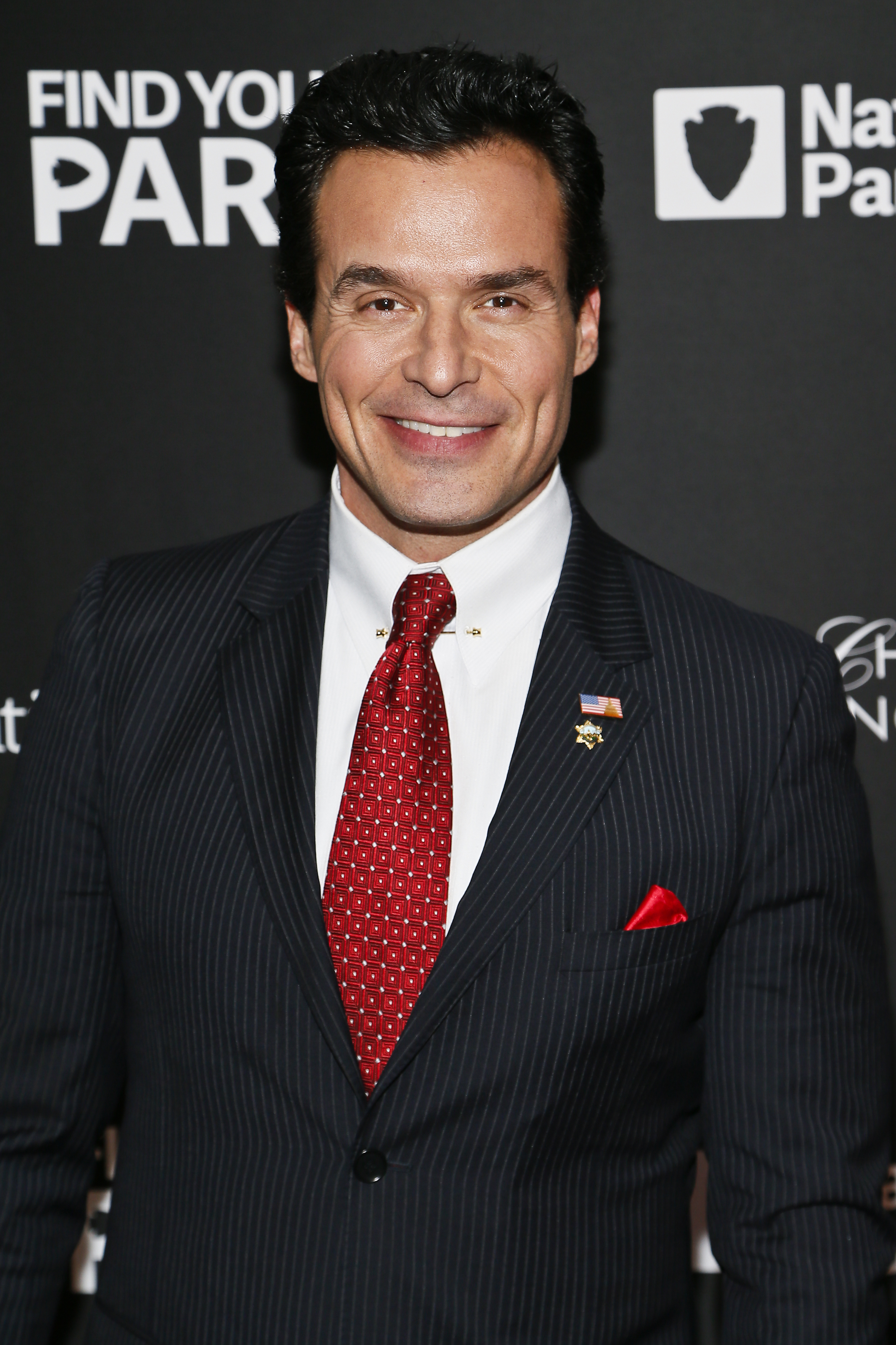 Antonio in pin-striped suit with patterned red tie and lapel pin smiles at a media event