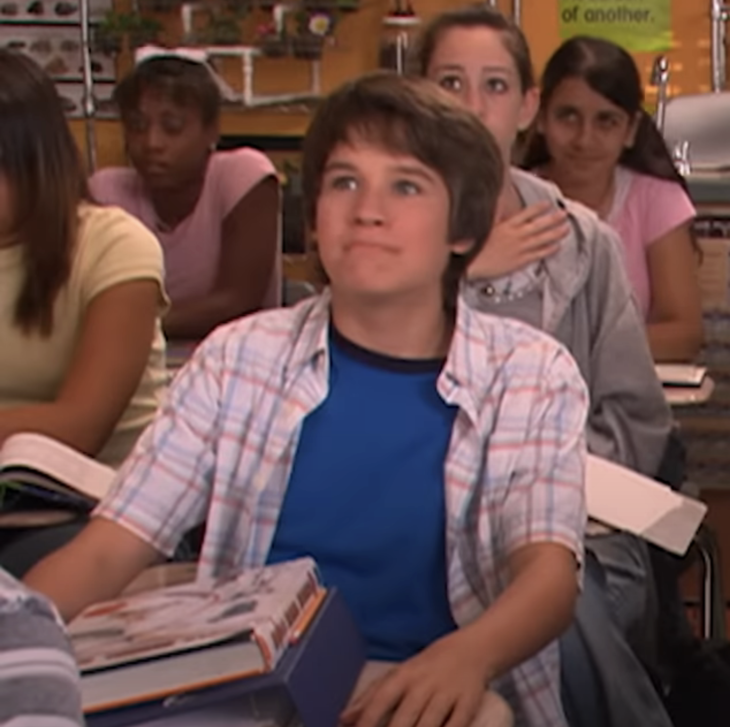 Devon as Ned wearing a plaid shirt over a tee, sitting in a classroom setting