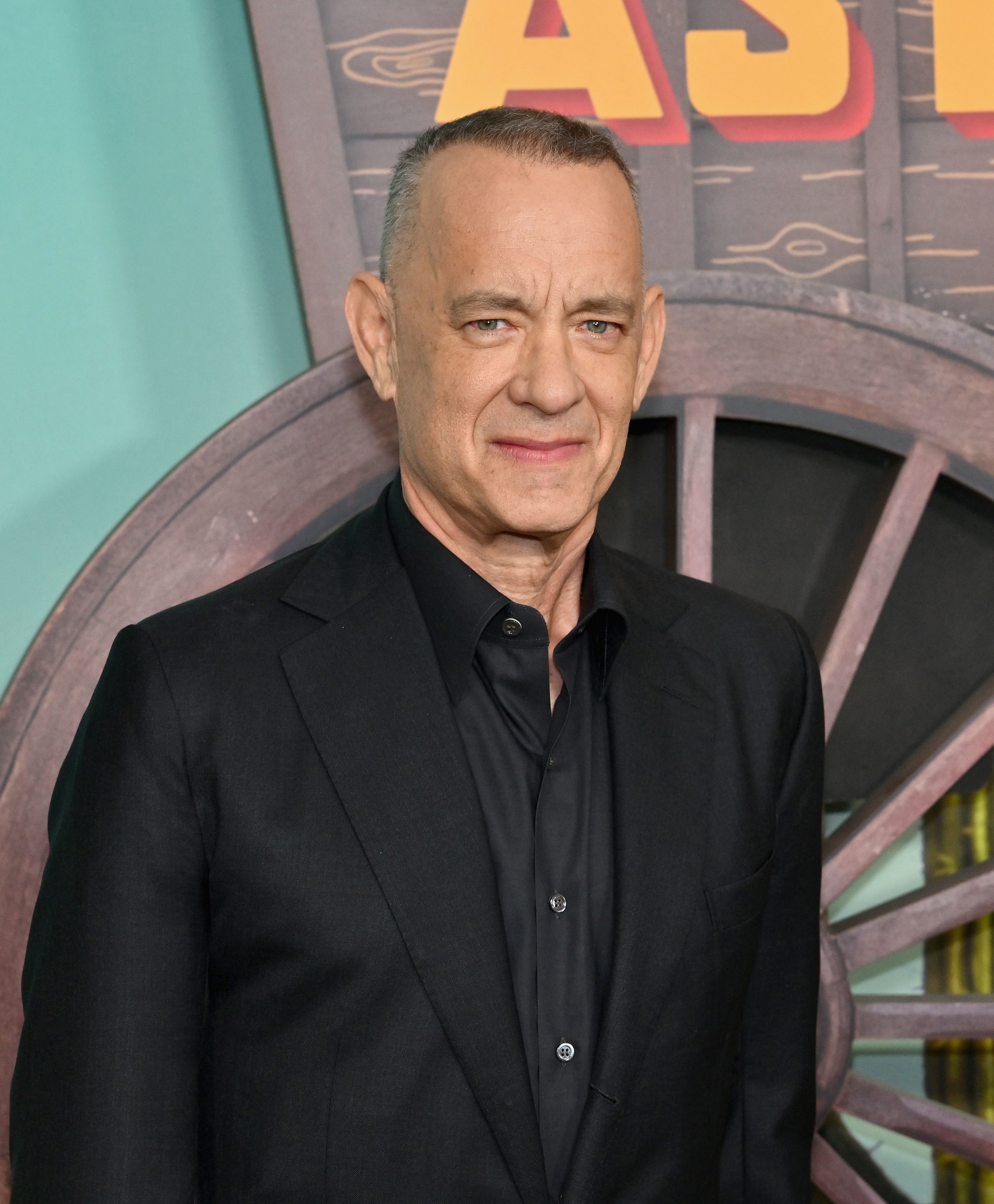 Tom Hanks wearing a classic black suit standing before a promotional backdrop