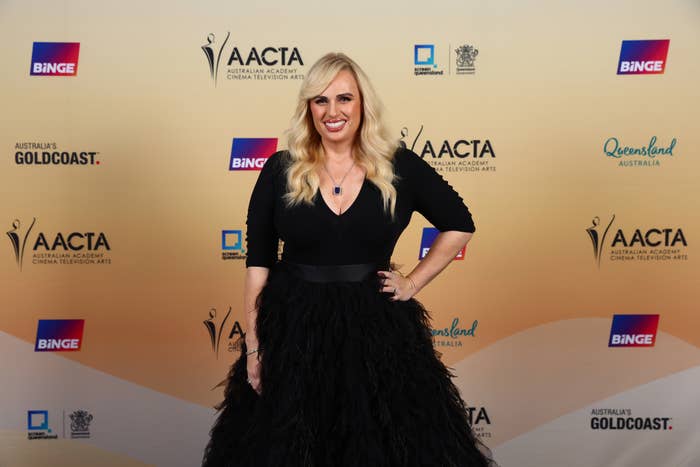 Rebel Wilson poses in a black dress with a fitted top and voluminous skirt at an awards event