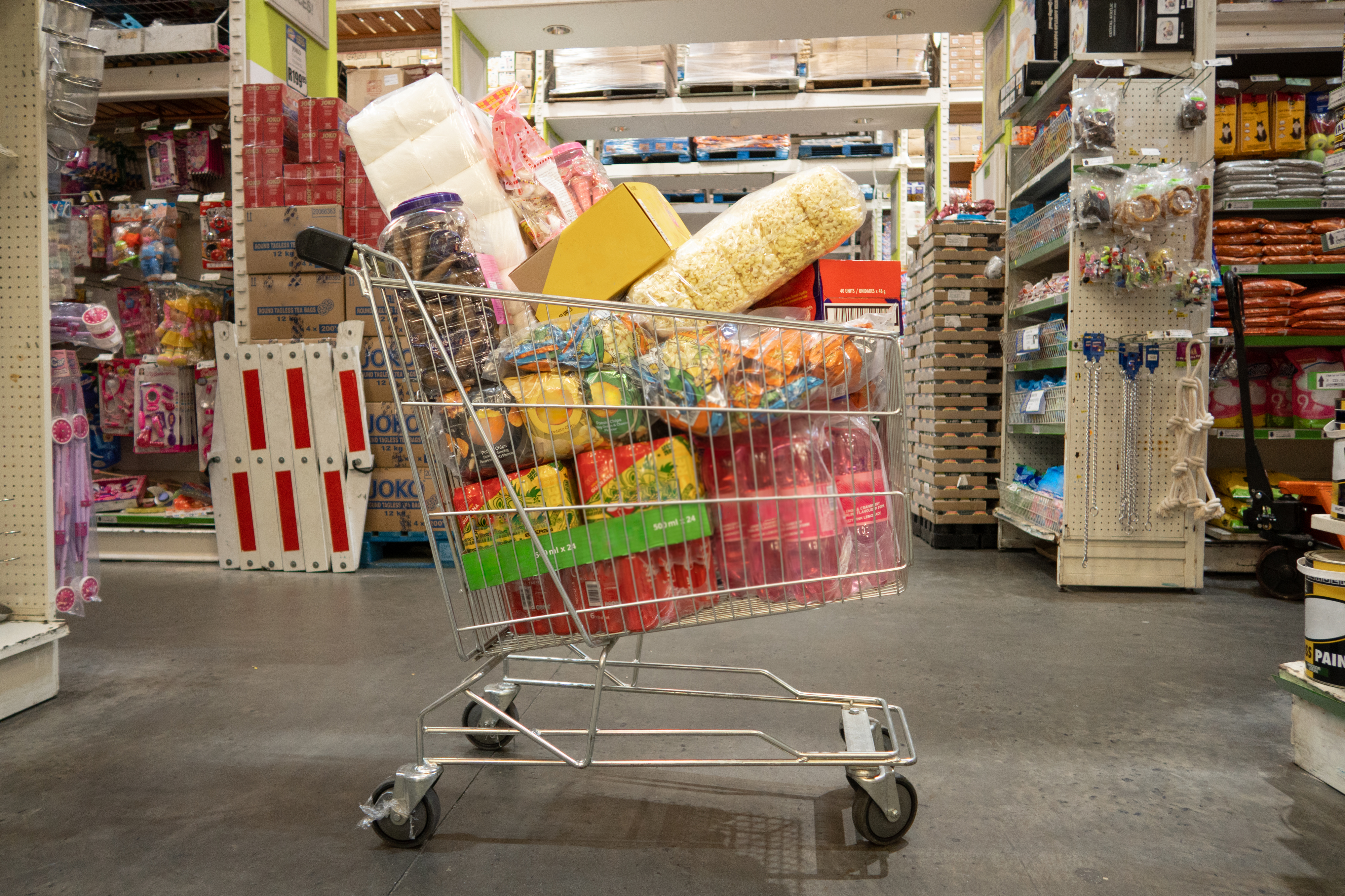 A shopping cart filled with various bulk sized household supplies and groceries inside a supermarket aisle