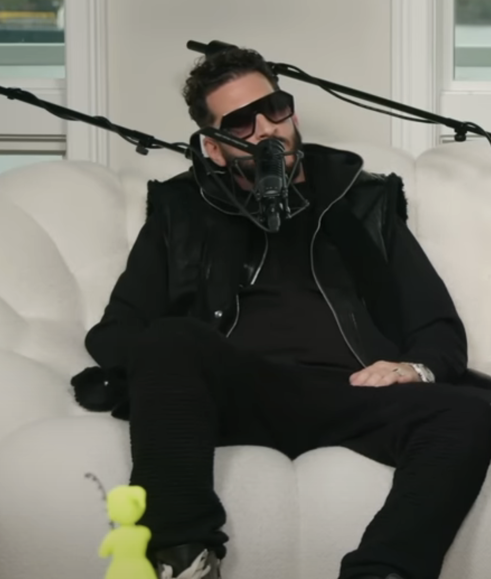 Jon in jacket and pants sits on a sofa with a microphone setup