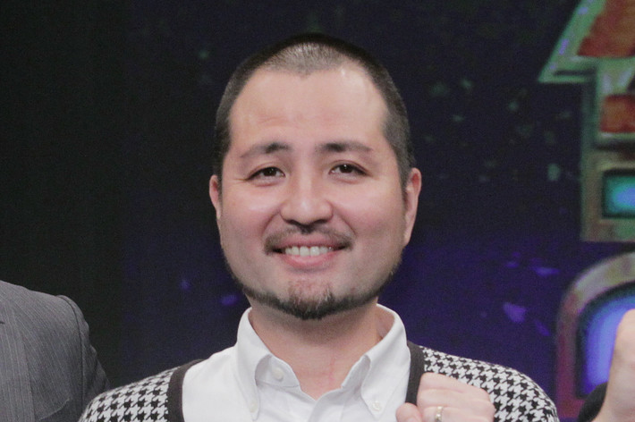 画像には、背景にキラキラした装飾があるステージ上に立つ複数の男性が映っています。会場はイベントのように見え、前列には男性が2人、それぞれ異なるポーズを取っています。