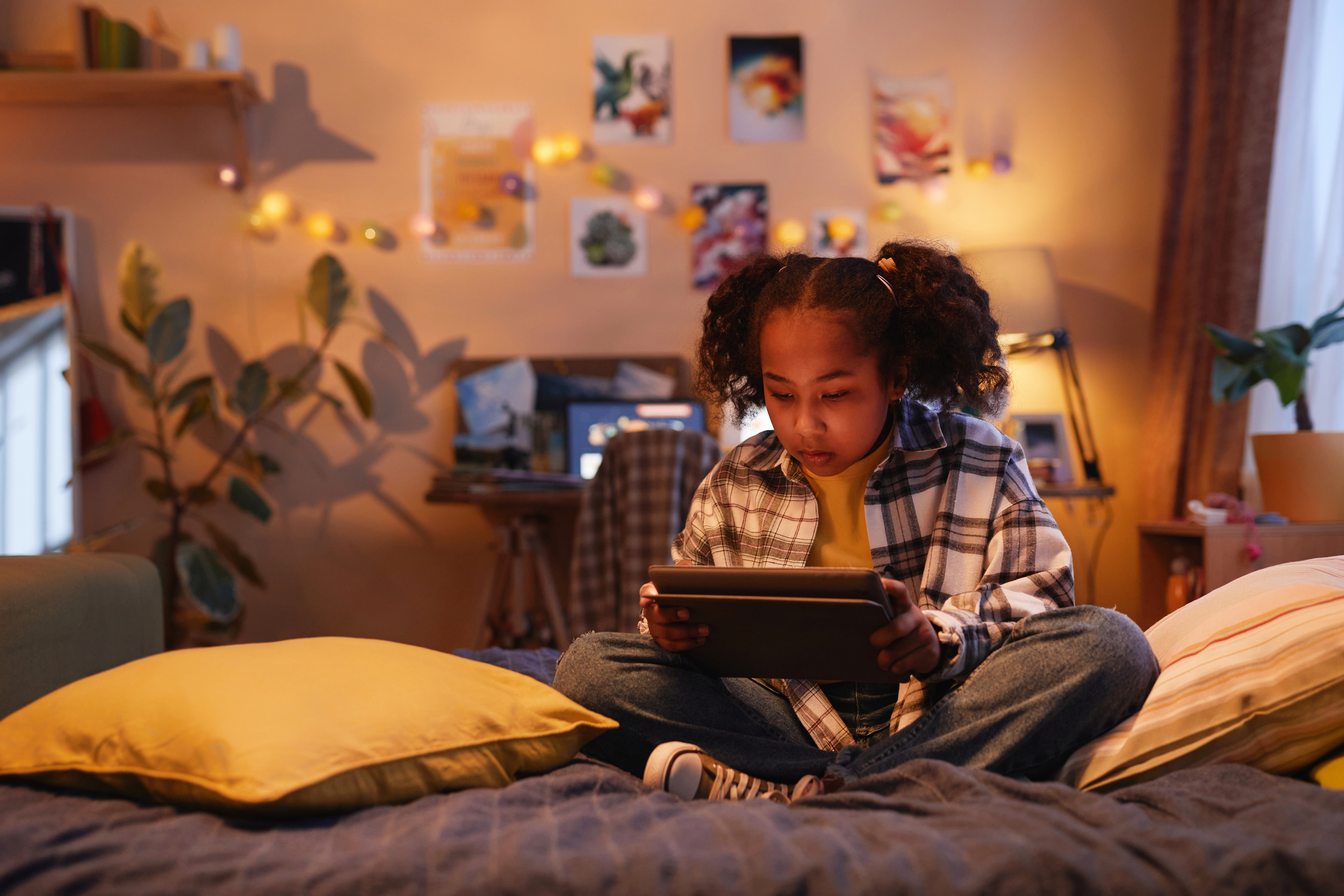 Child sitting on bed engaged with a tablet, surrounded by pillows, with artwork on the wall
