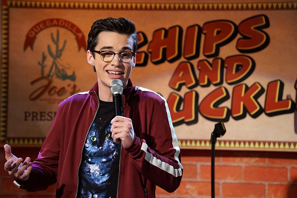 Joey in a scene on stage holding a microphone with graphic tee and jacket, in front of a sign reading &quot;Chips and Chuckles&quot;