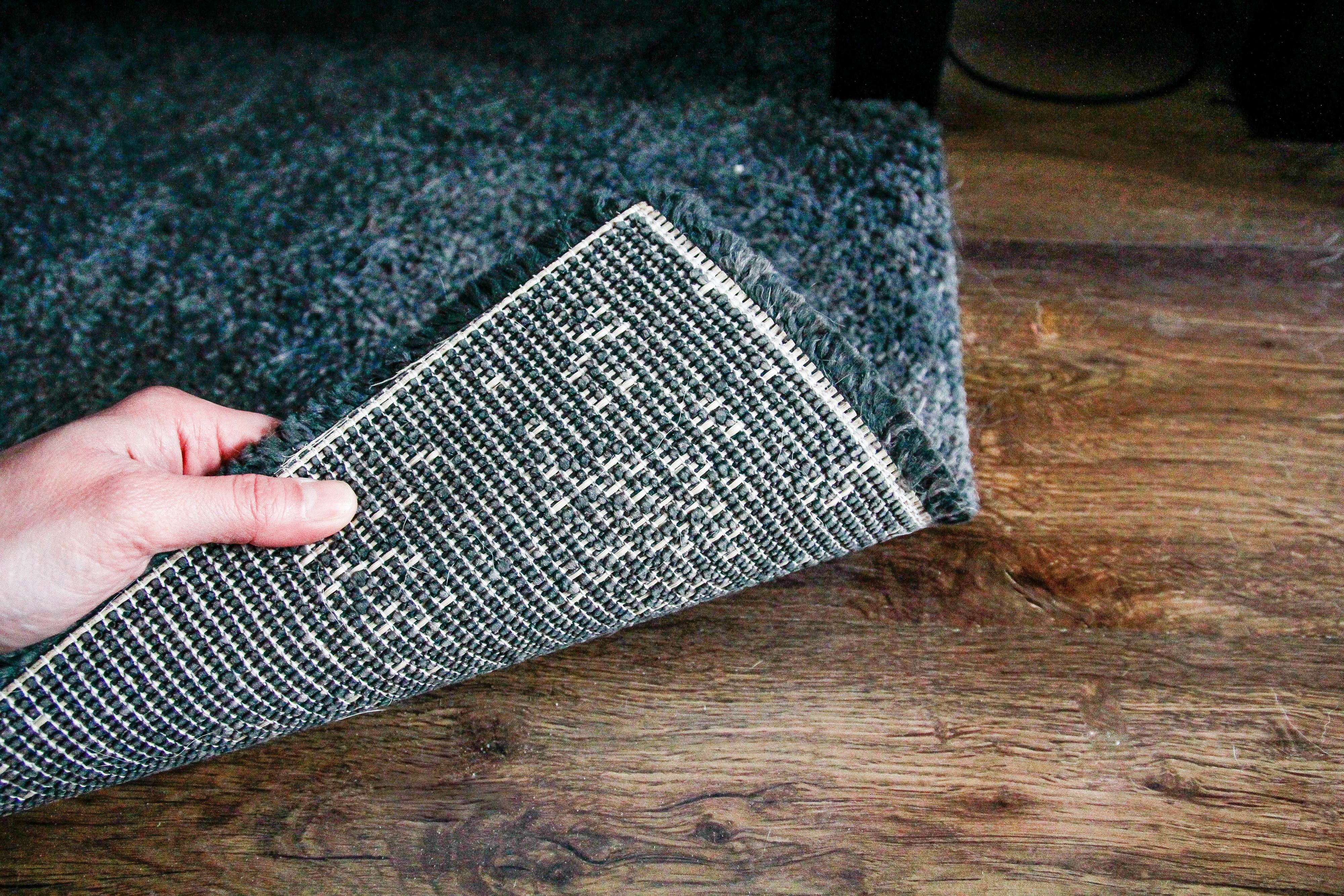 Person lifting corner of a textured rug to reveal wooden flooring underneath