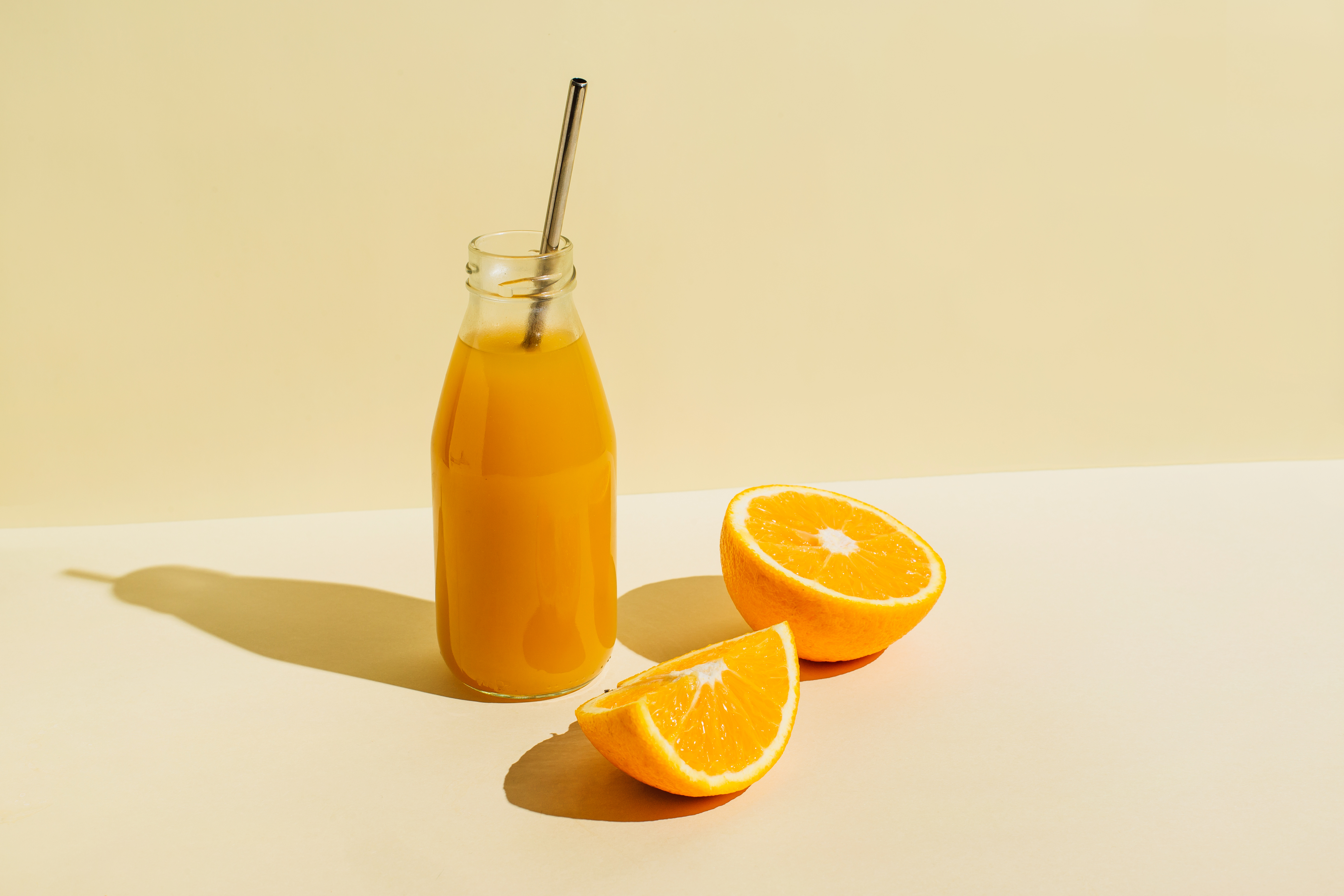 Bottle filled with an orange beverage with a metal straw next to halved oranges on a surface
