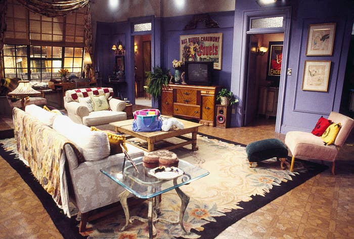 Set from TV show with sofa, chairs, and glass coffee table in a living room. Various eclectic decorations are visible