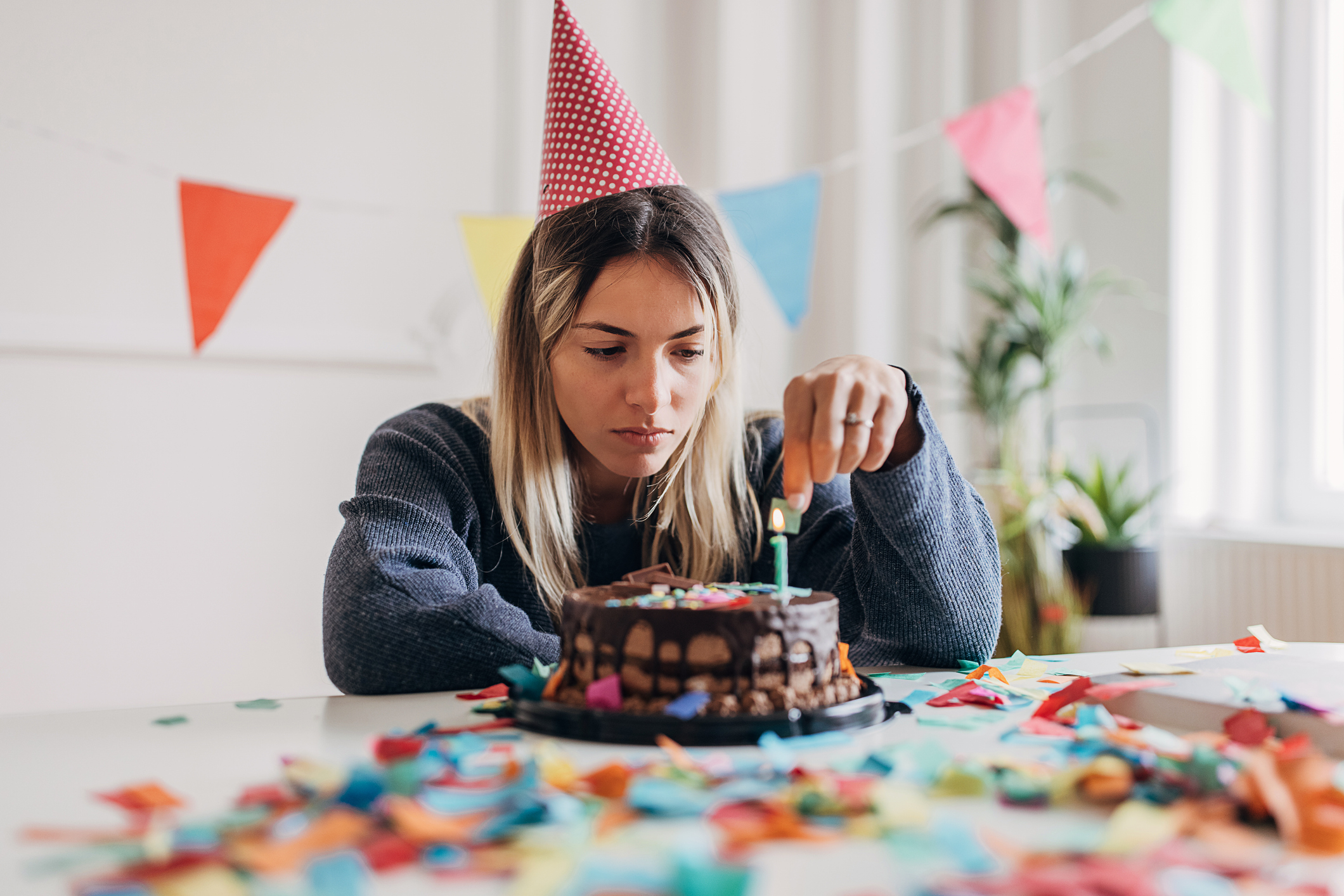 Sad-looking young woman at a table looks at a birthday cake with candles, party hats and confetti around