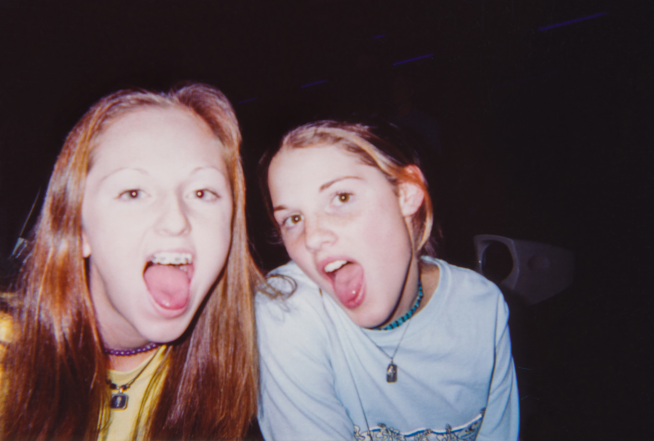 Two girls sticking out their tongues, wearing casual tops and beaded necklaces