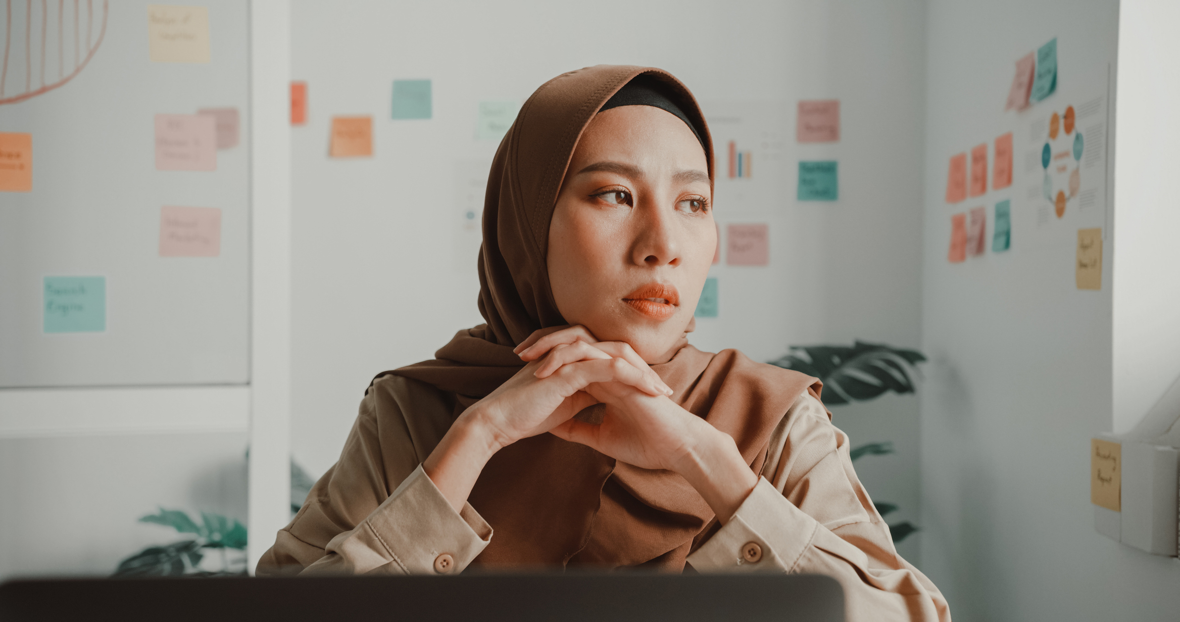 Woman in hijab looking contemplative at desk with sticky notes in background
