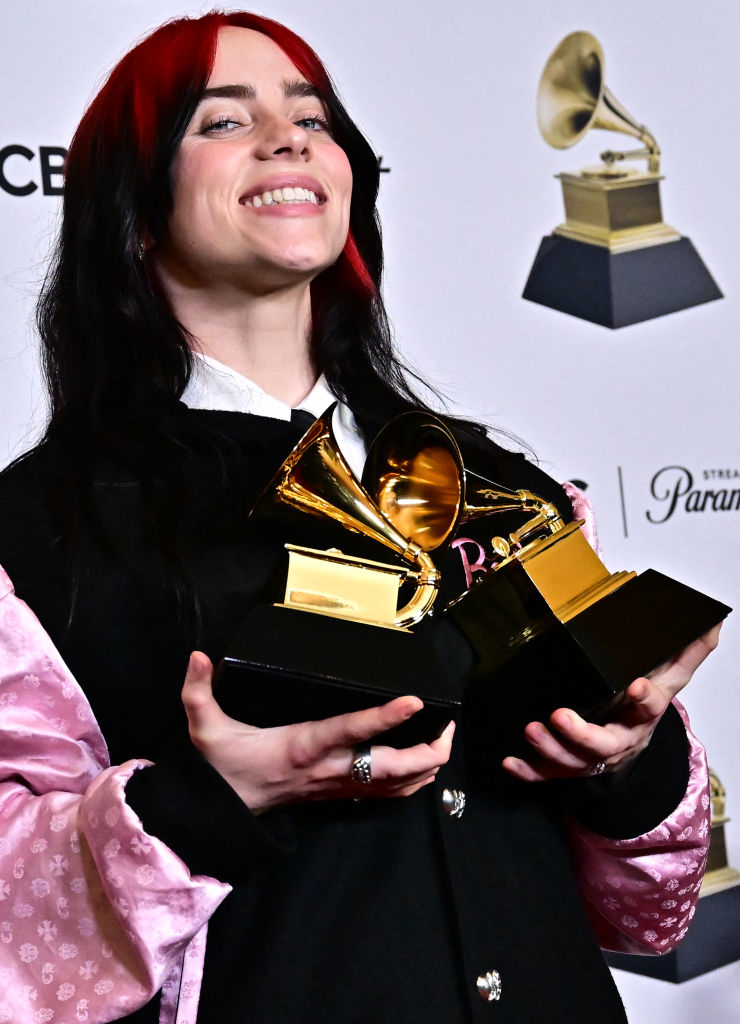 Billie Eilish smiling, holding a Grammy Award, wearing a black and pink outfit