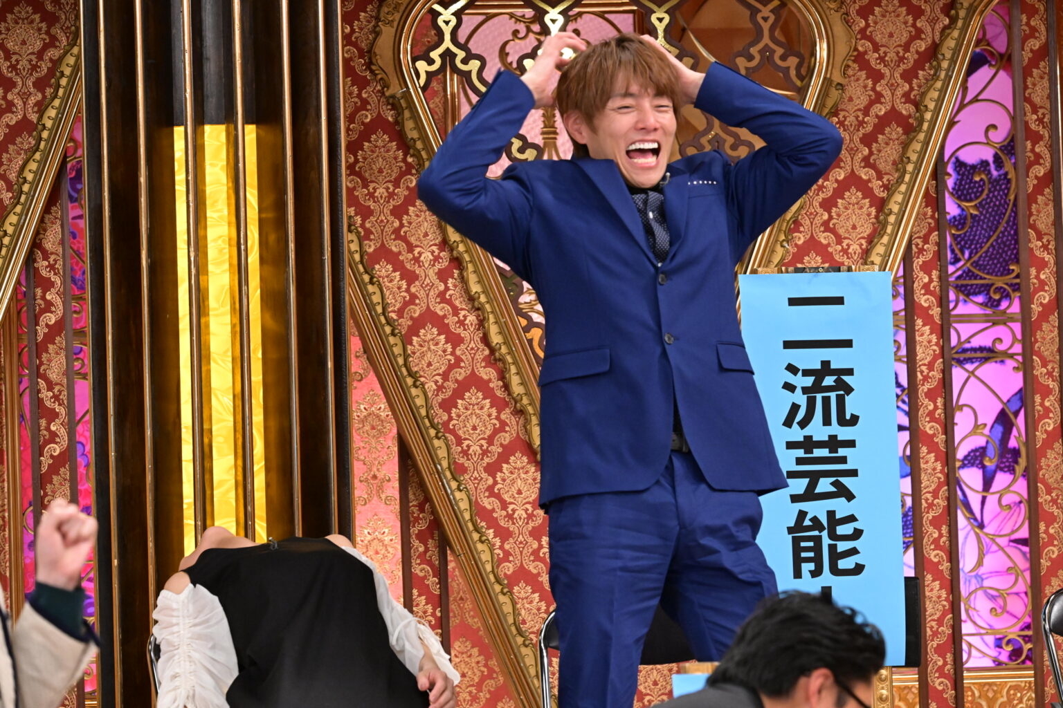 男性がステージ上で青いスーツを着て喜びのポーズをとっている。