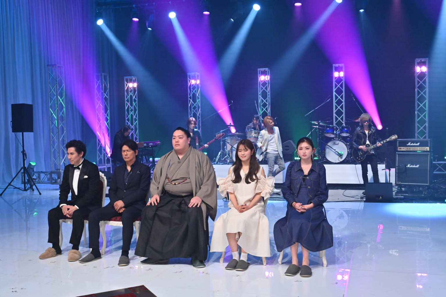 ステージ上で着物を着た男性とドレス姿の女性が並んで座っていて、バンドセットが背後にあります。