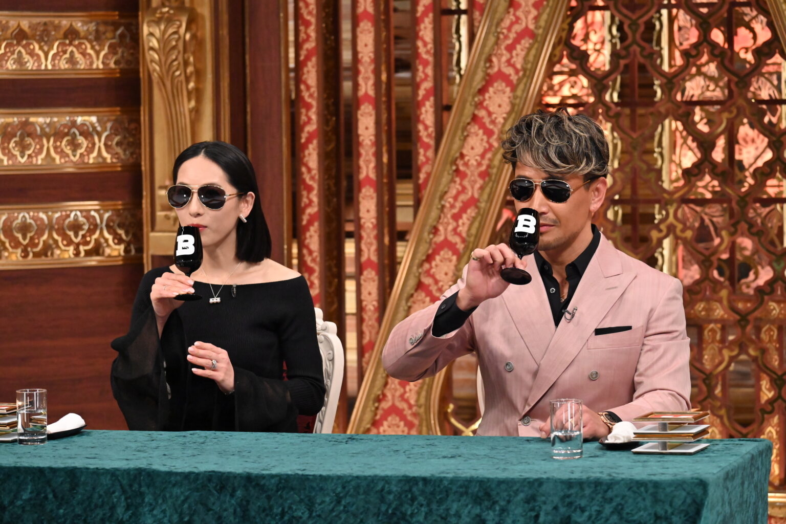 二人の有名人がマイクを持ってテーブルに座っている。右の人物はピンクのスーツ、左は黒いドレス。