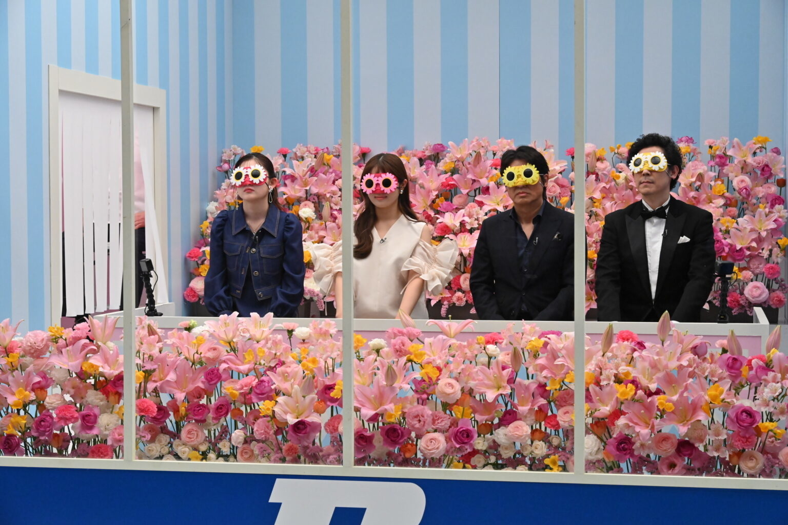 四人が花に囲まれた展示スペースに立って、顔に花の形のメガネをかけている。