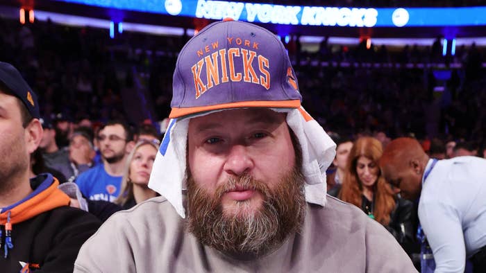 Man in Knicks hat at basketball game, focused on camera with crowd in background