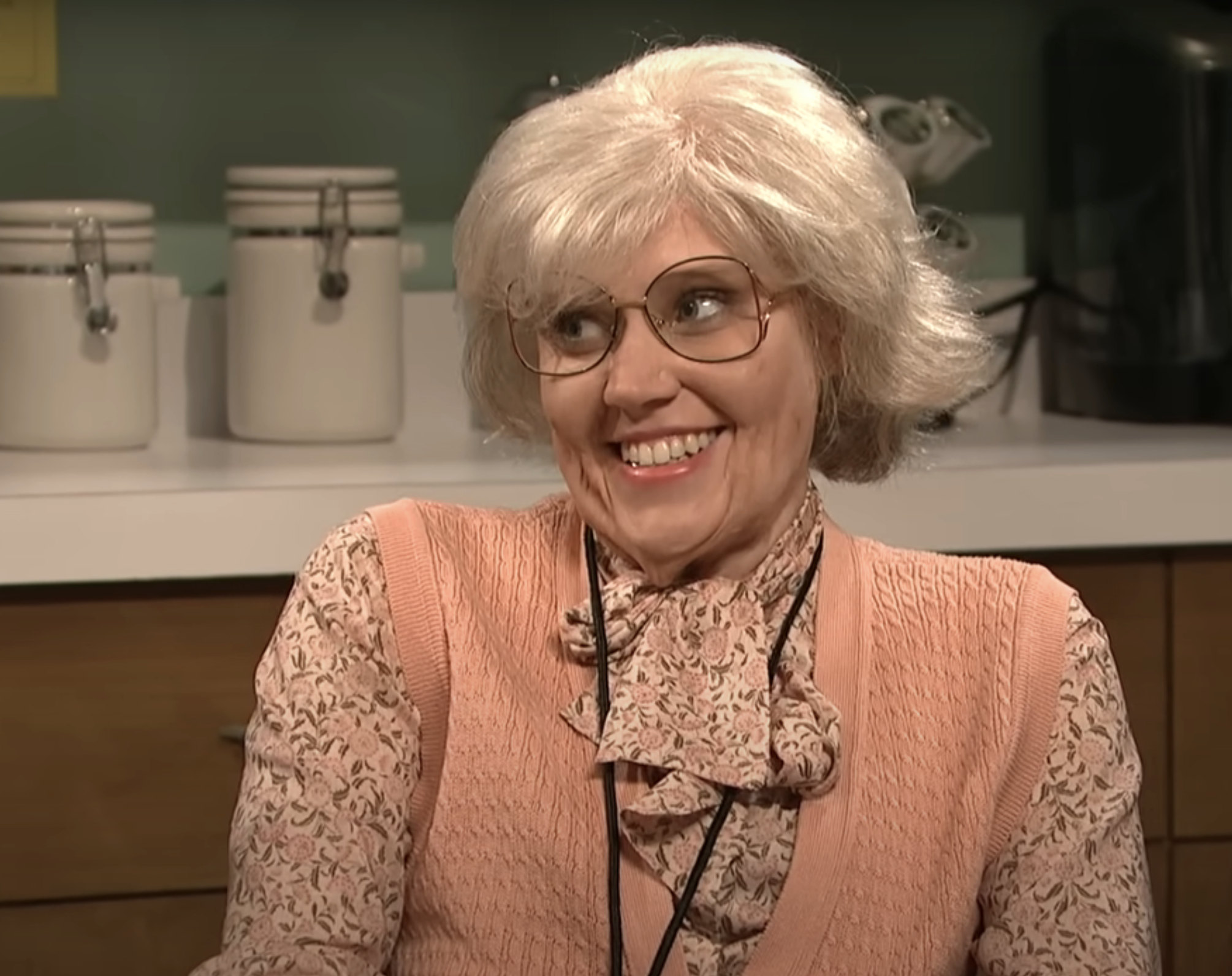 Kate McKinnon as an older lady with glasses wearing a floral shirt, sweater vest, and pearl necklace in a kitchen set