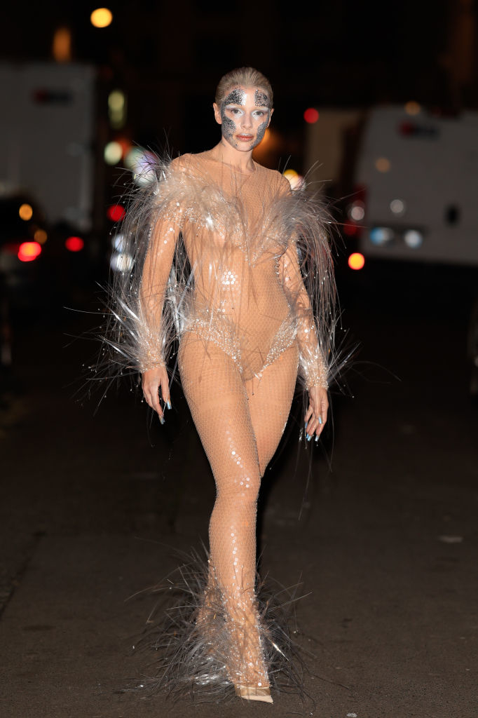 her in a sheer, embellished bodysuit with feather-like adornments, walking at a fashion event