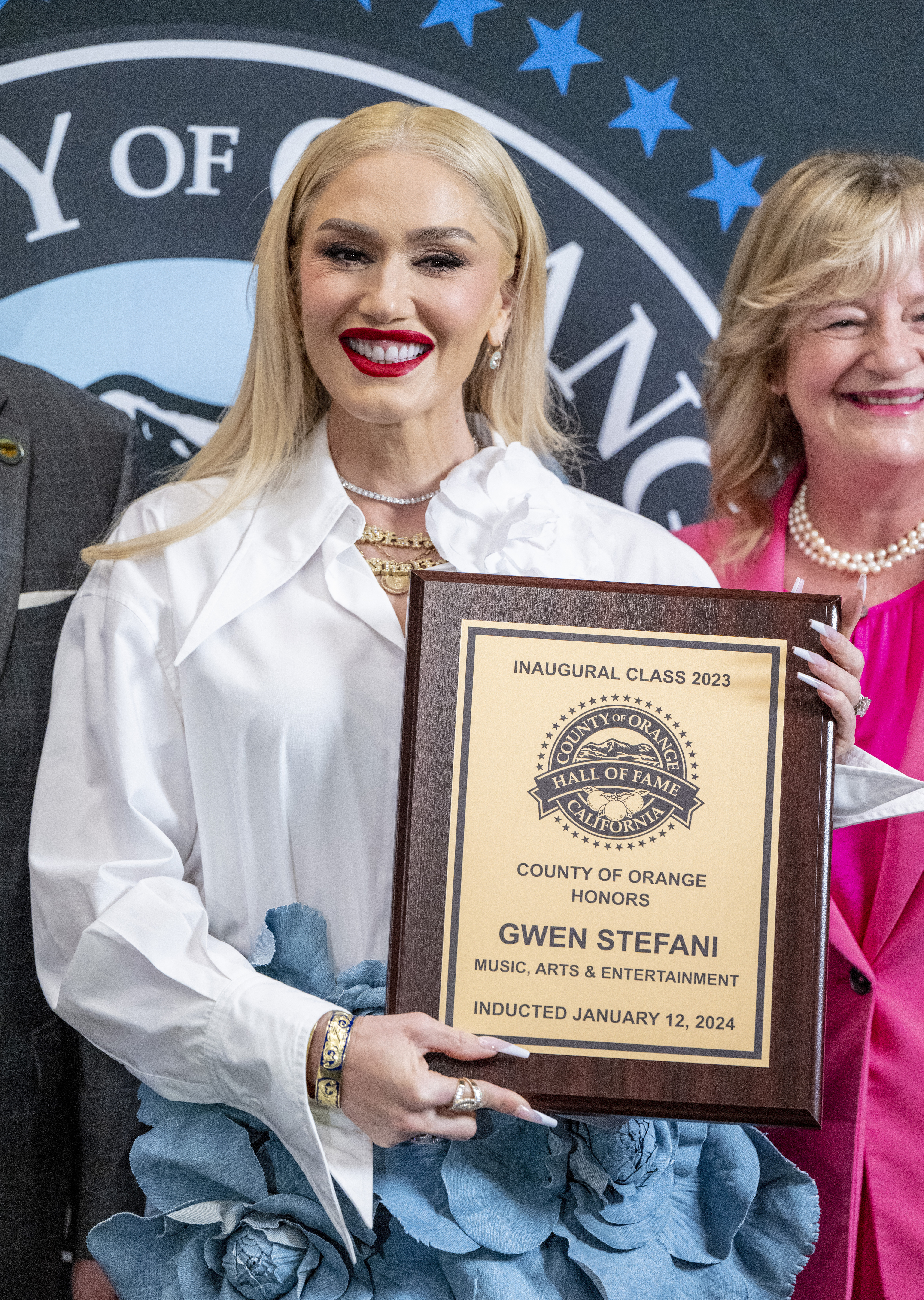 Gwen Stefani holding a plaque at the Orange County Music, Arts &amp;amp; Entertainment Hall of Fame 2023 event, smiling, with two individuals partly visible behind her