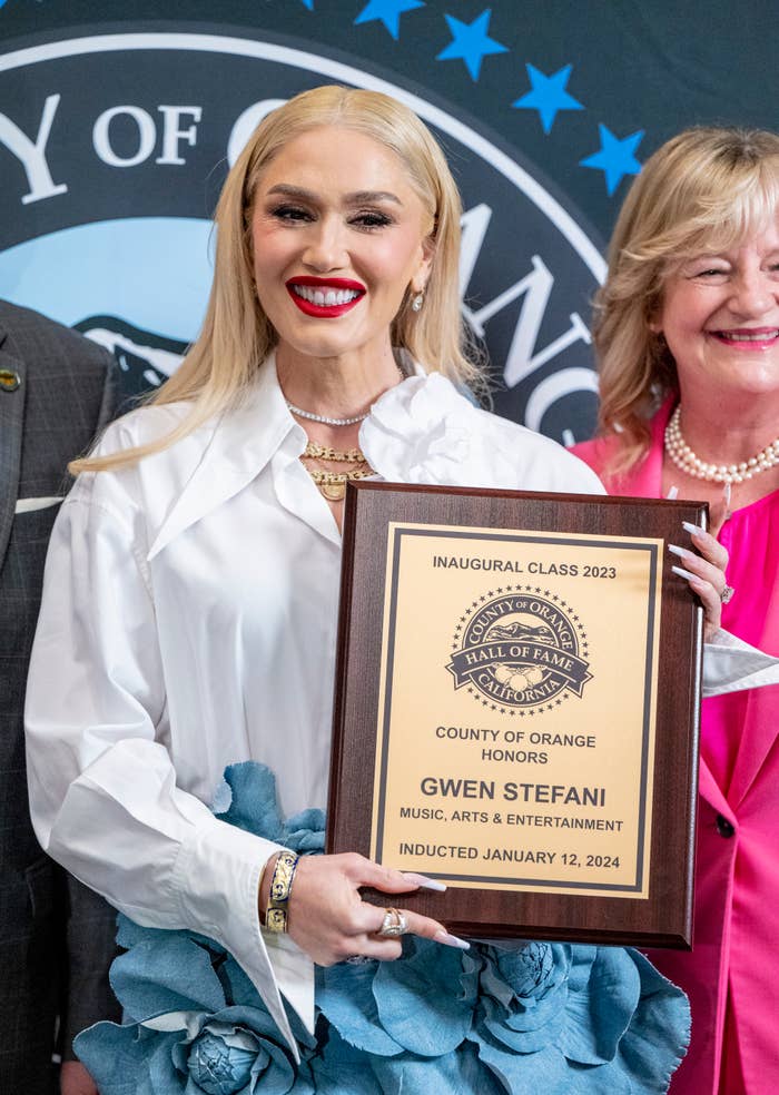 Gwen Stefani holding a plaque at the Orange County Music, Arts &amp; Entertainment Hall of Fame 2023 event, smiling, with two individuals partly visible behind her