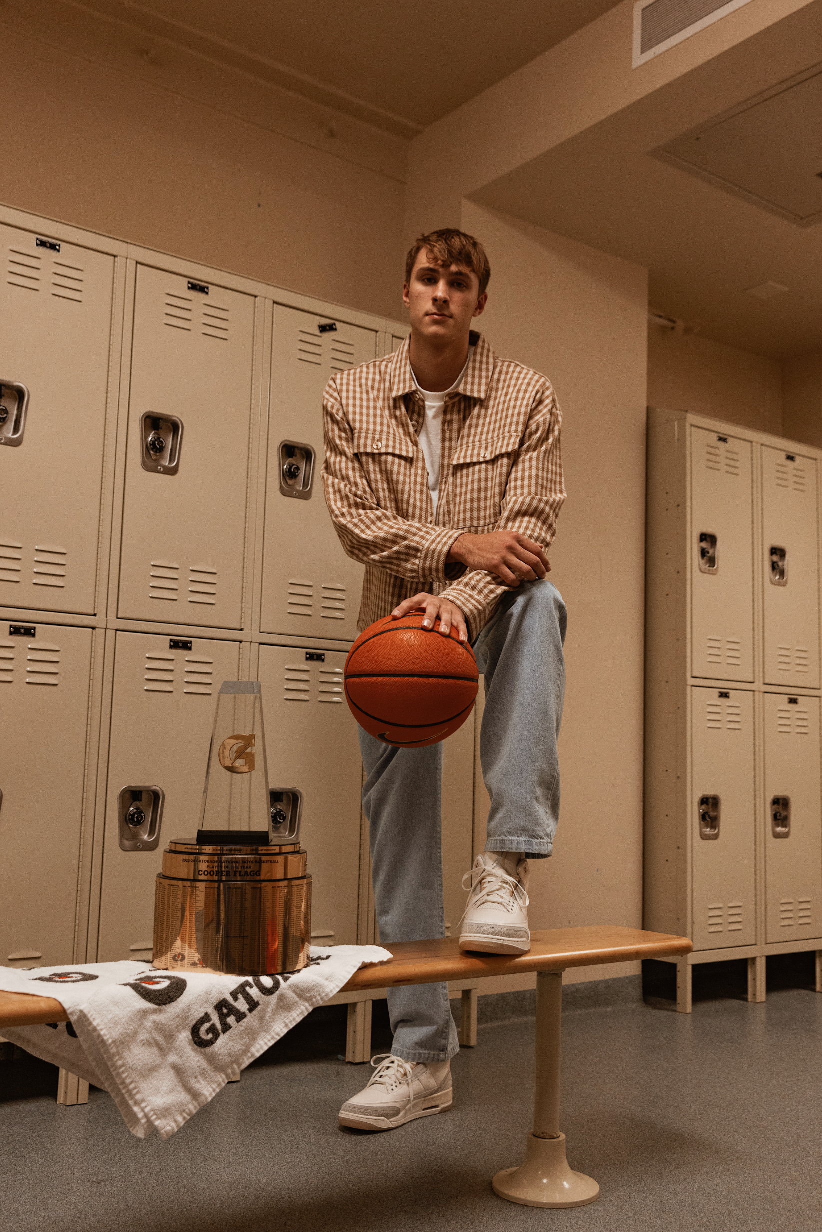 Person in a locker room sitting with a Basketball, wearing plaid jacket and jeans