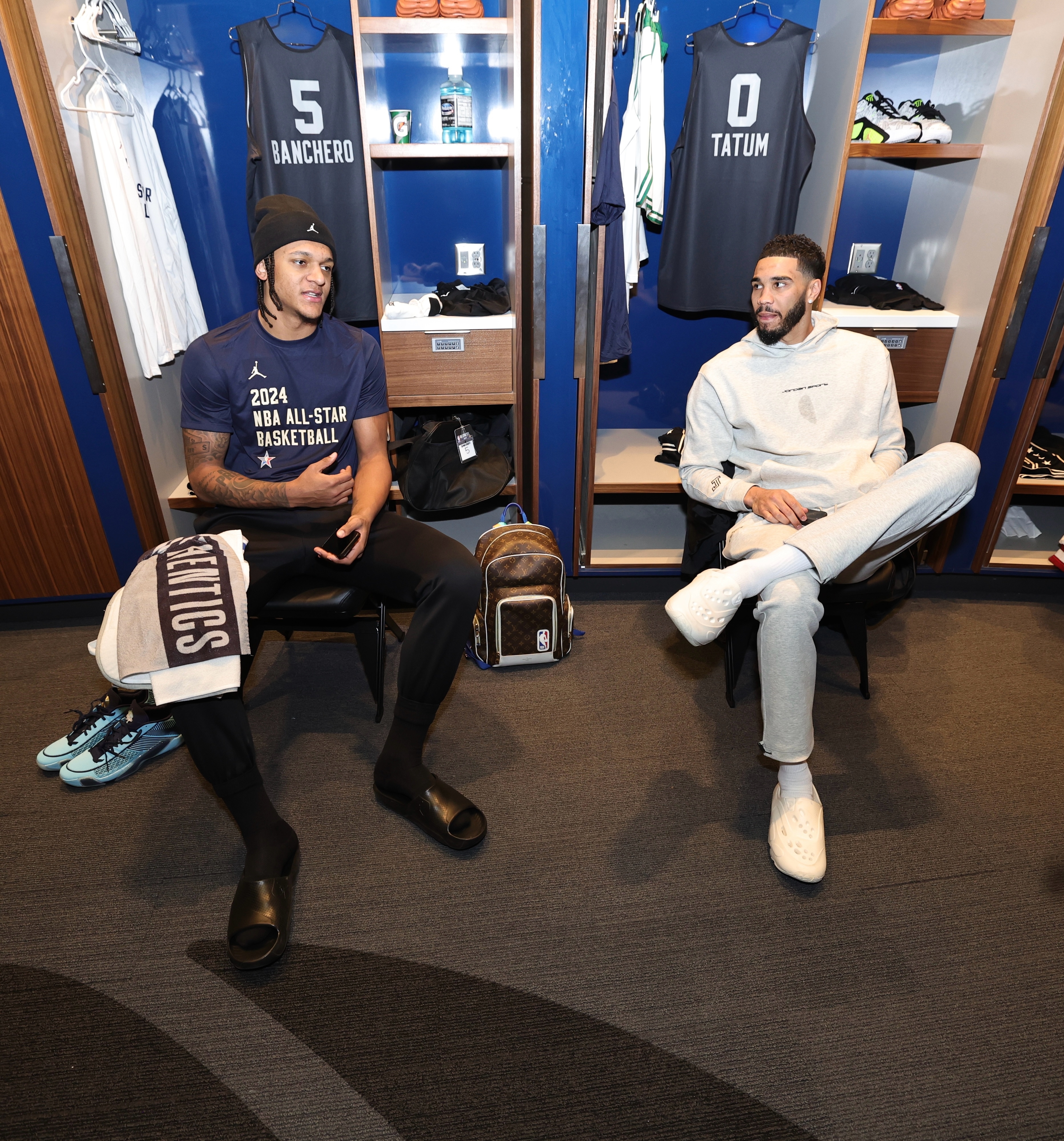 Two athletes sitting in a locker room, one in a navy NBA All-Star shirt, the other in a gray sweat outfit