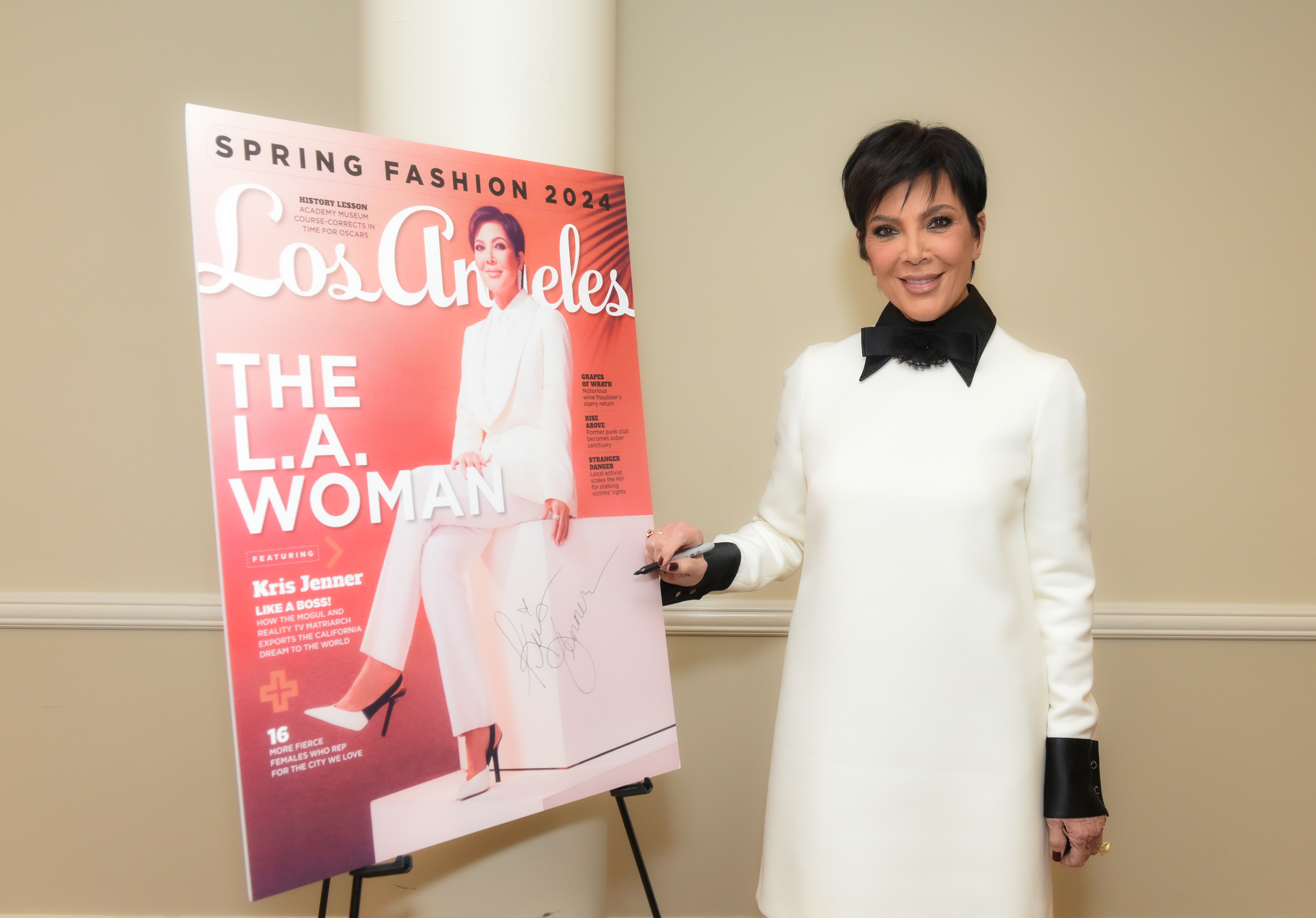Kris Jenner stands beside a magazine cover featuring her, wearing a white dress with a black bow tie neckline