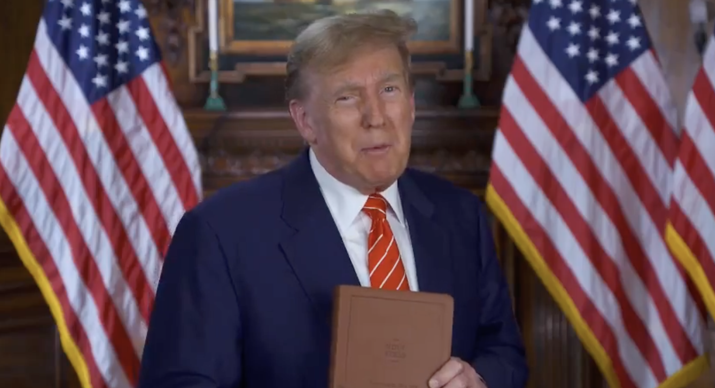 Donald Trump stands with a book, flanked by two US flags, addressing the camera