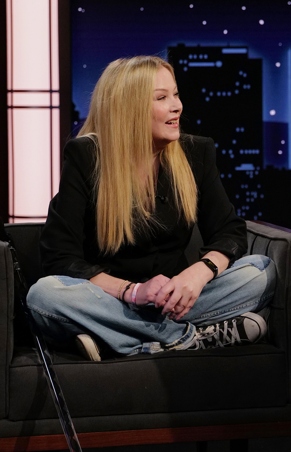 Celebrity sitting cross-legged on a couch during a talk show, wearing a black blazer and denim jeans