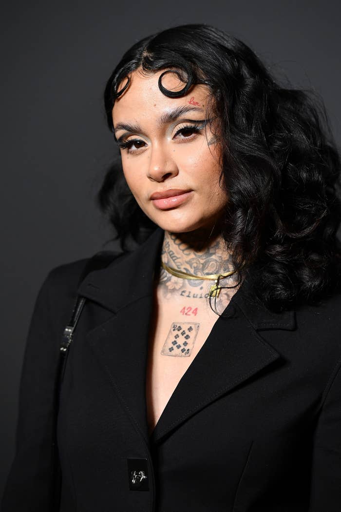 Kehlani with curled hair, facial tattoos, wearing a dark blazer with a pin