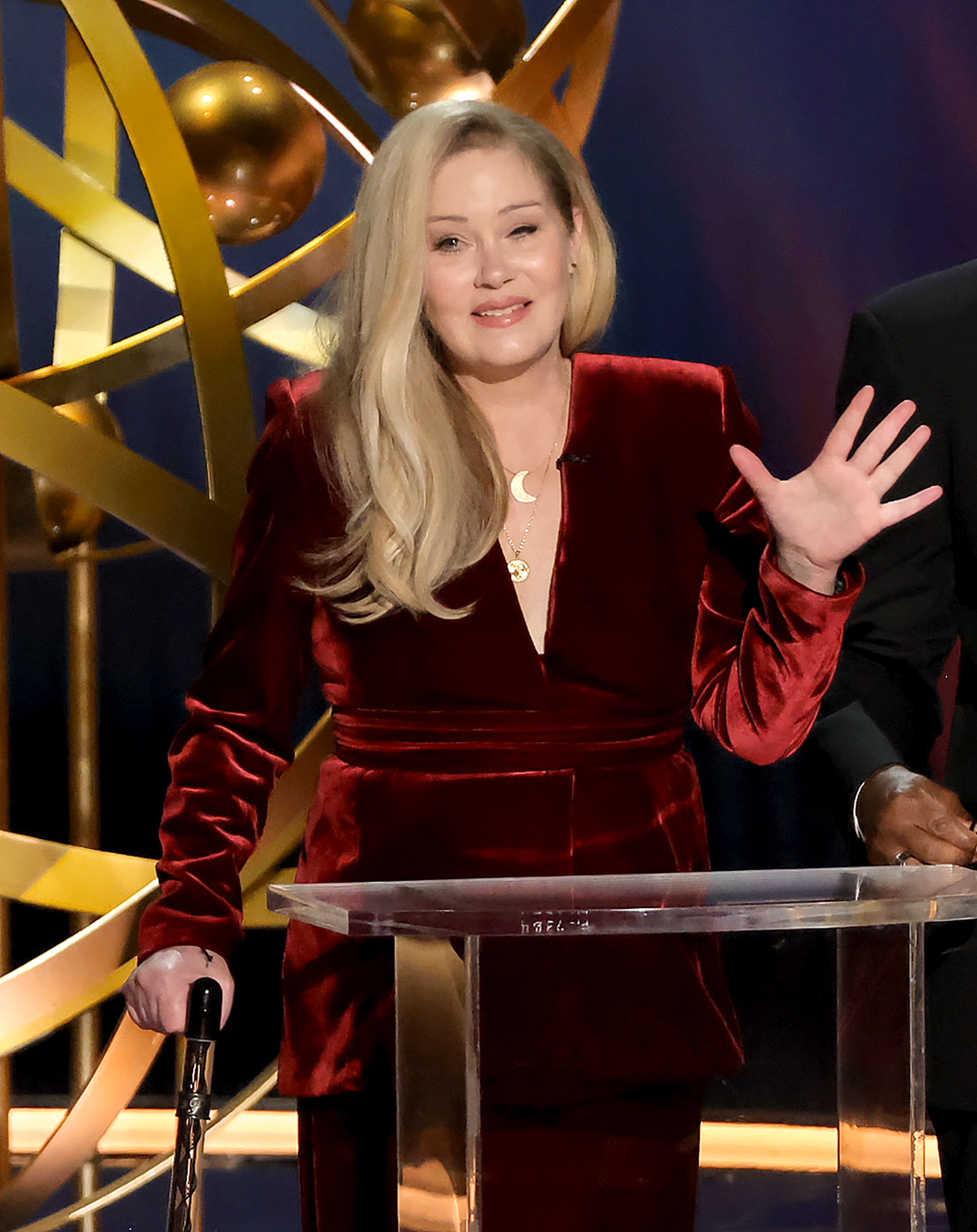 Christina Applegate speaking at a podium, wearing a velvet suit, waving to the audience