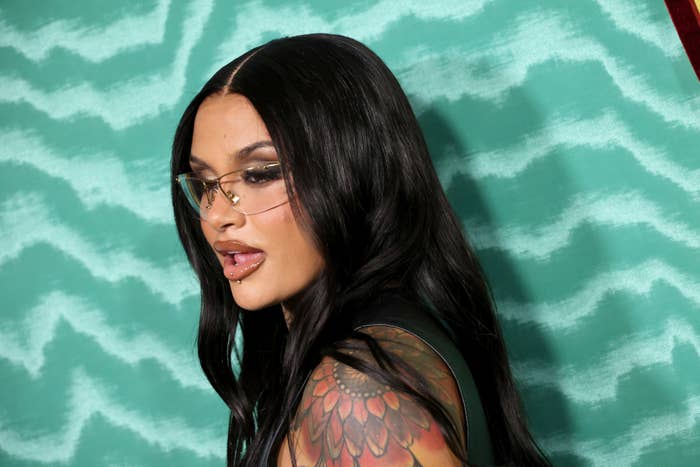 Kehlani with long hair and glasses, tattooed shoulders, in front of a wavy patterned backdrop
