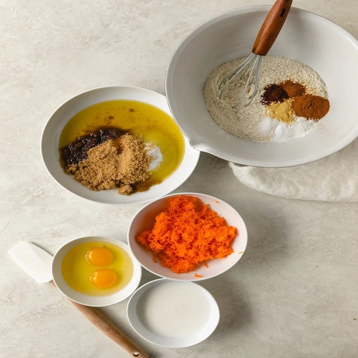 Ingredients for baking are separated into bowls: eggs, flour mixture, shredded carrots, and sugars with oil