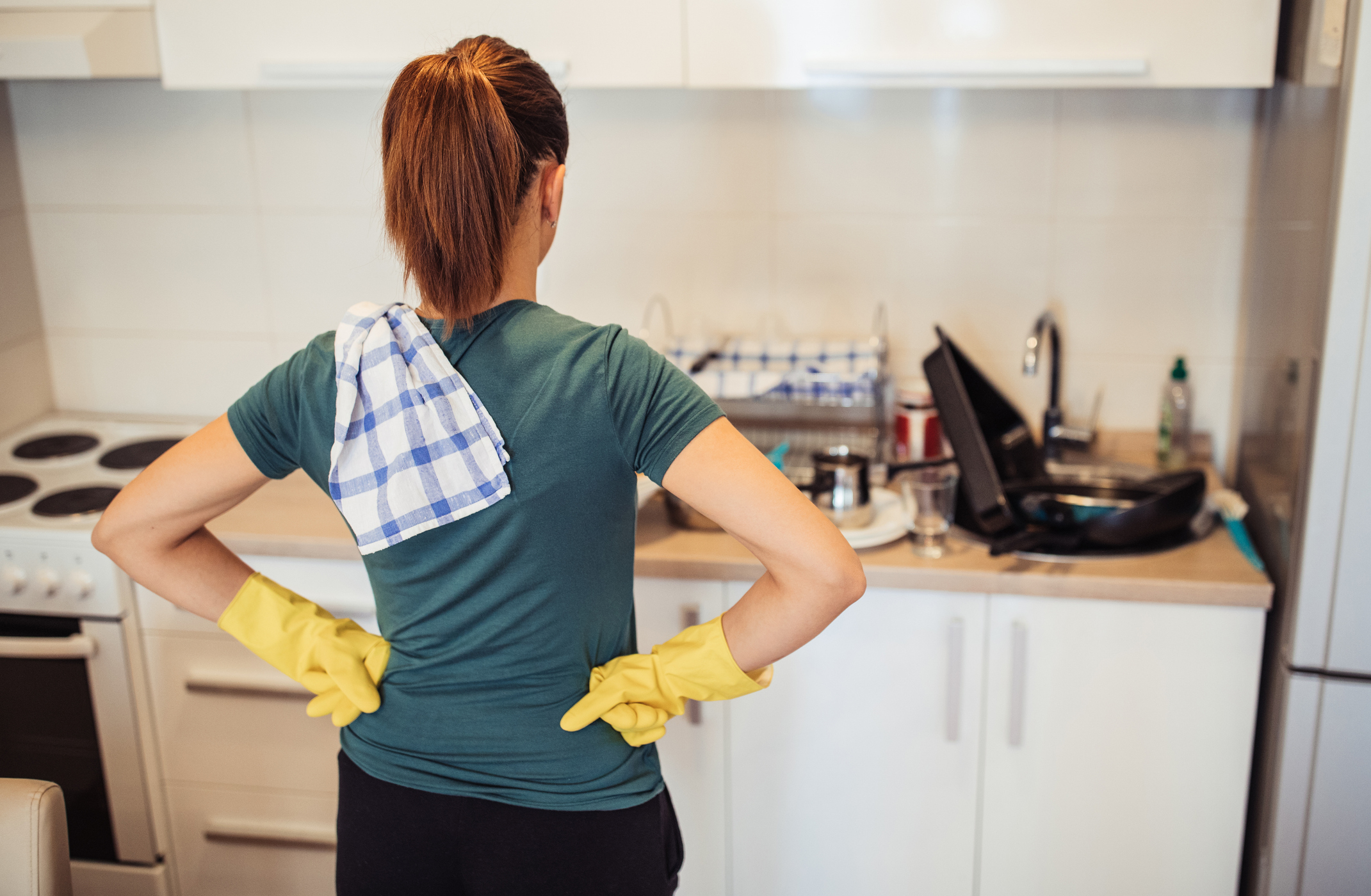 Woman with rubber gloves stands with hands on hips facing dirty kitchen, ready to clean