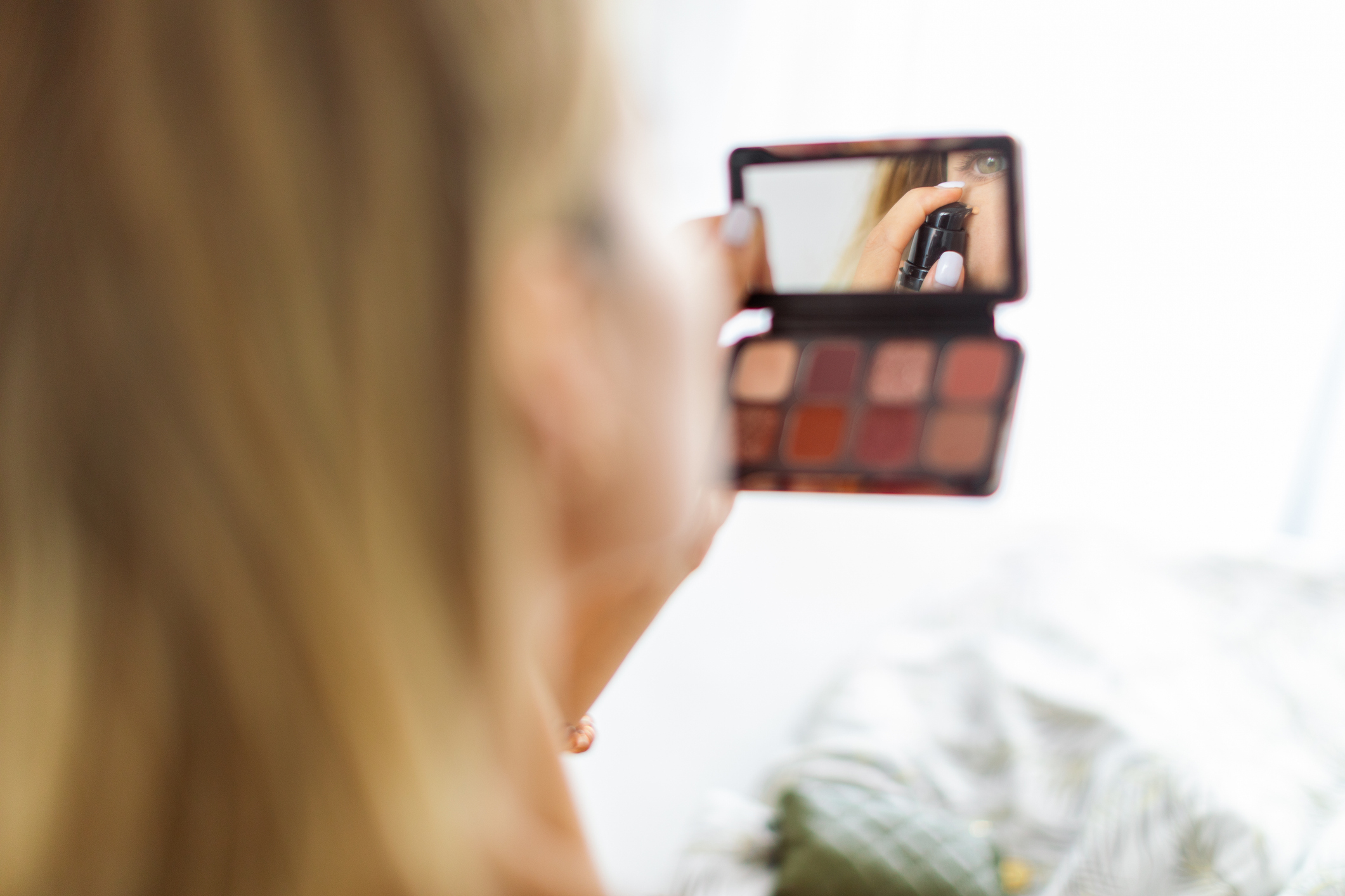 Woman looking into a handheld makeup palette mirror, applying eye shadow. (Personal names are not provided in the image.)