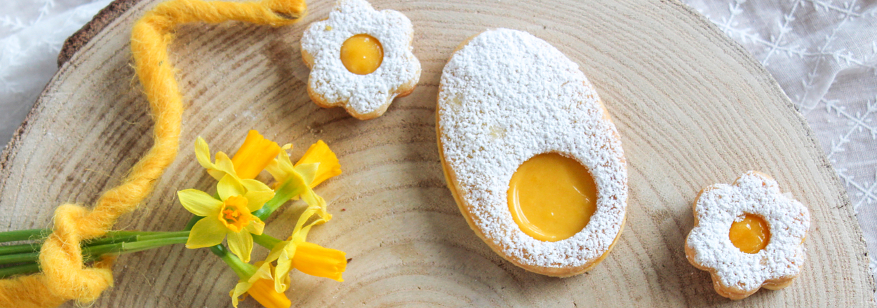 Easter cookies shaped like eggs and flowers with lemon curd centers on a wooden surface next to yellow flowers