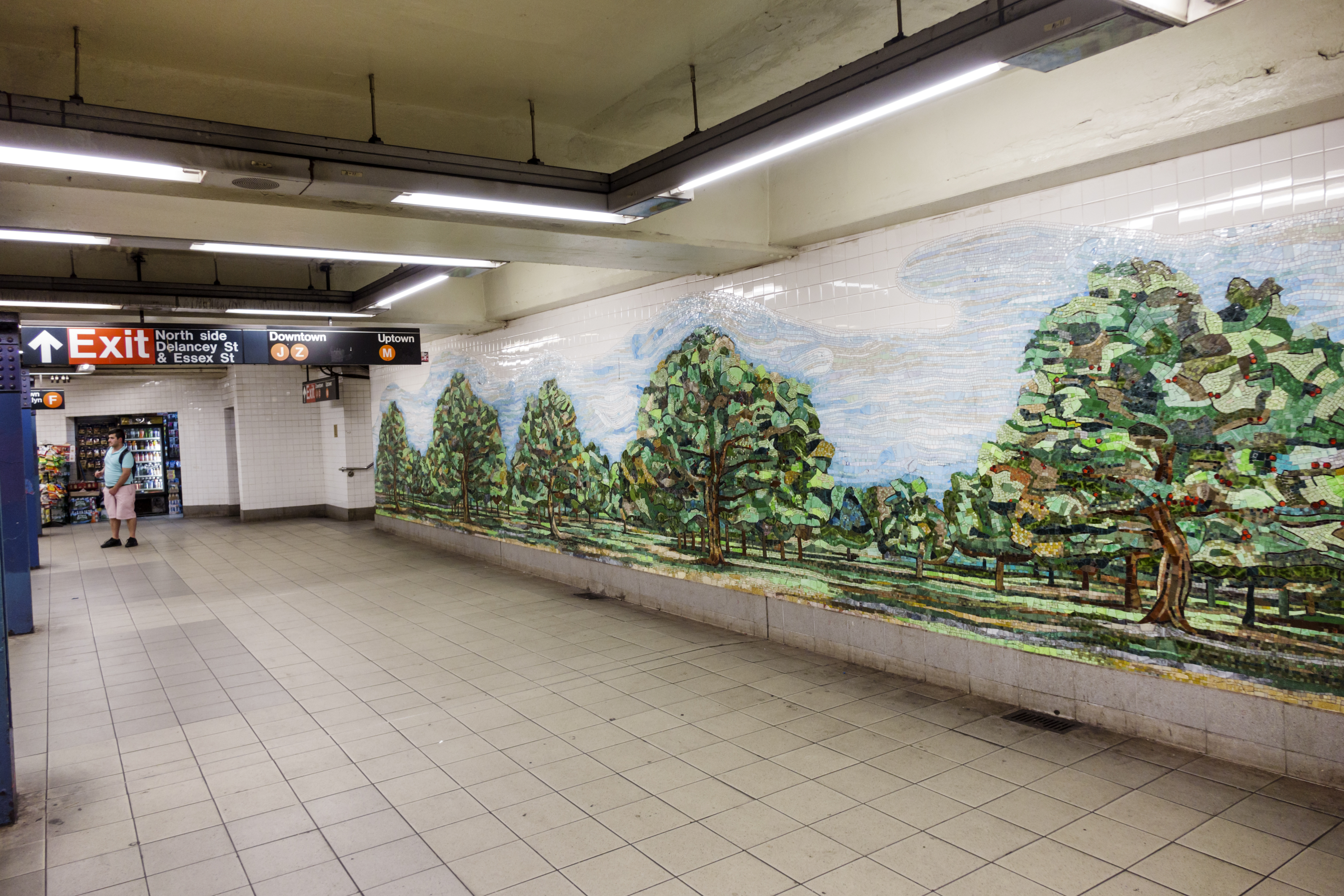 Person stands in a subway station with a mosaic artwork of trees on the wall. Exit sign visible