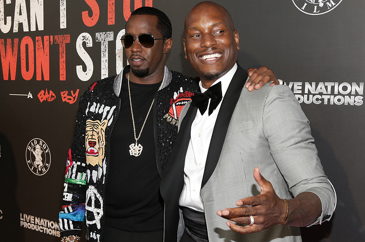 Two men on a red carpet, one in a graphic tee and blazer, the other in a suit and bow tie, posing for the camera