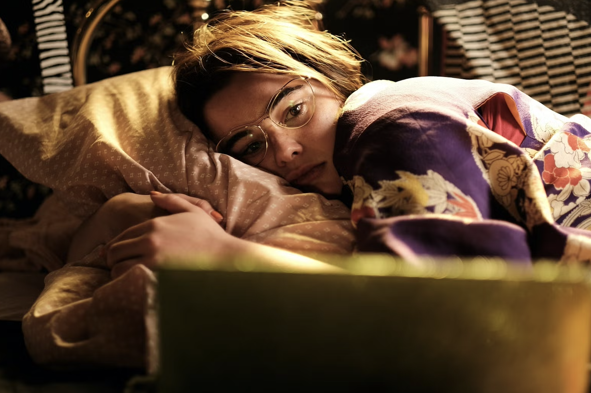 Person resting on a couch, wrapped in a blanket, wearing glasses, looking at laptop.