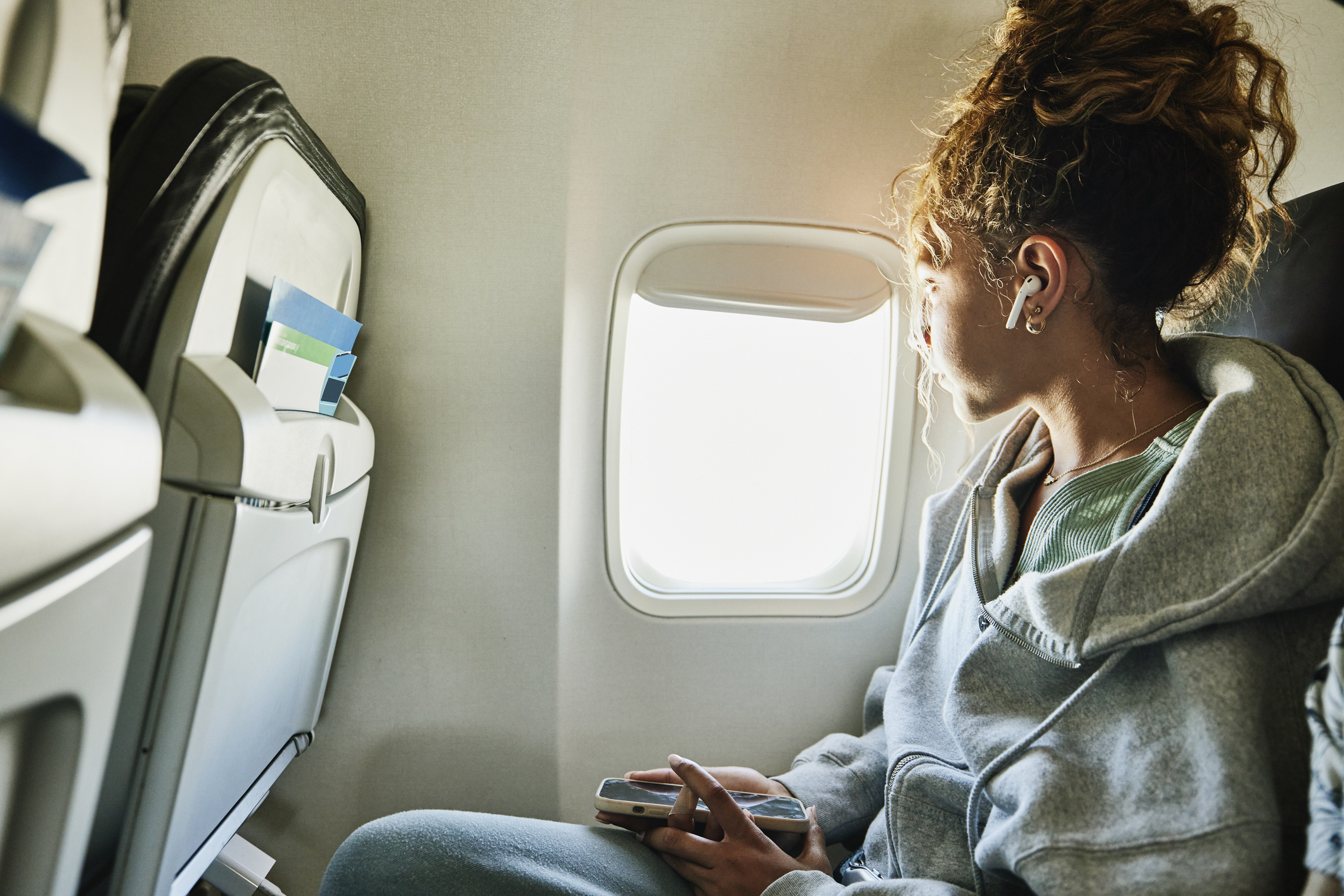 Person with headphones looking out an airplane window, holding a phone, with a seatback visible