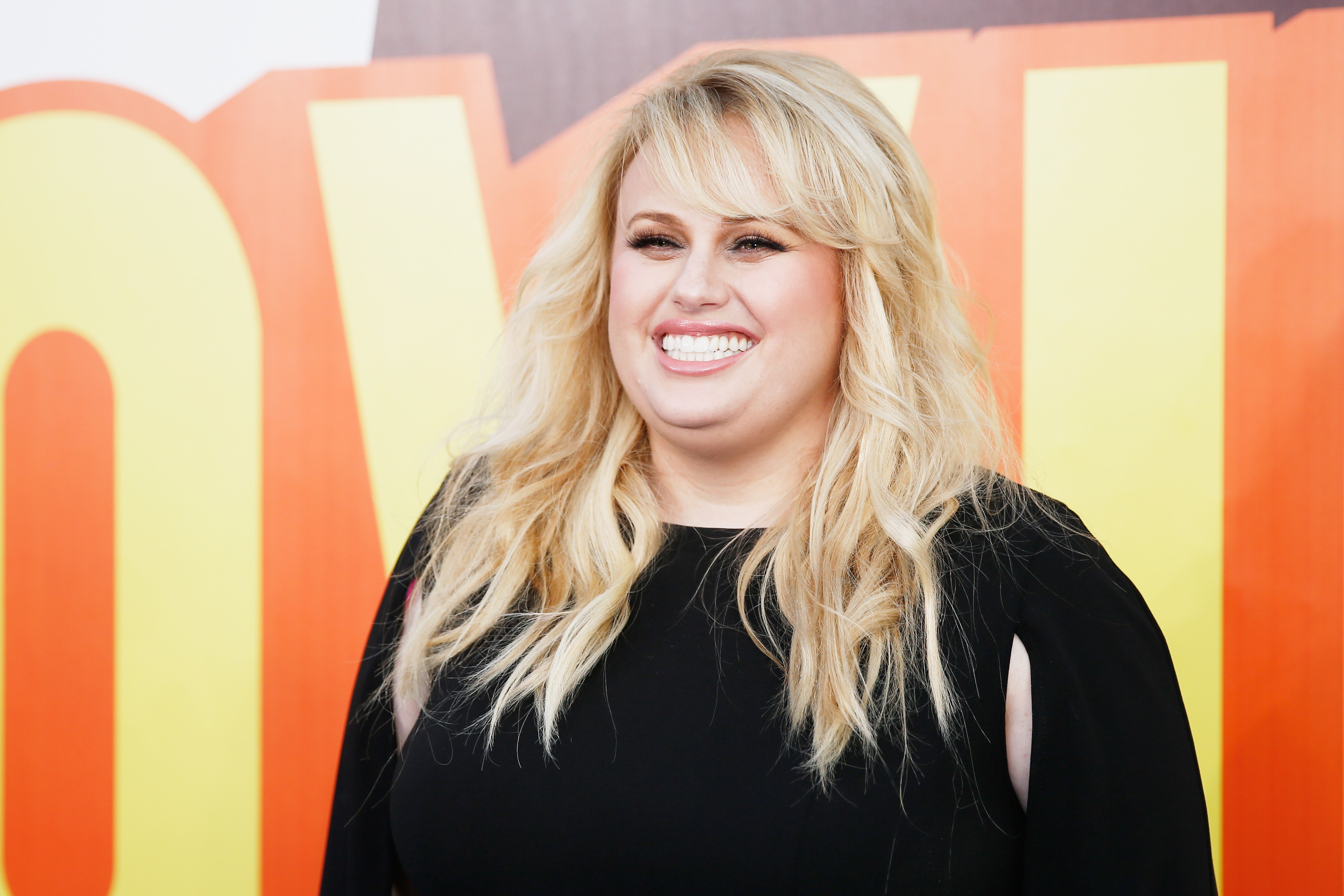 Rebel Wilson smiling at an event in a long-sleeved  dress with ] sleeves