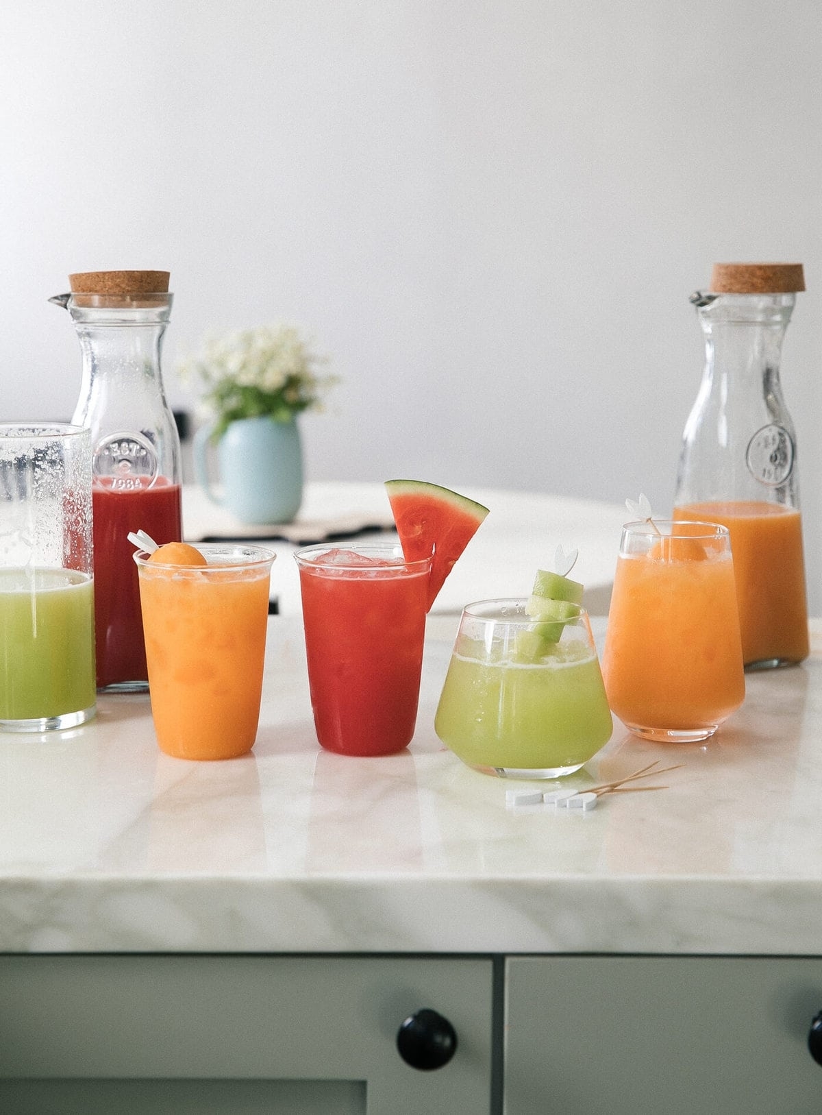 Assorted fresh juices in clear glasses on a kitchen counter, with two bottles in the background
