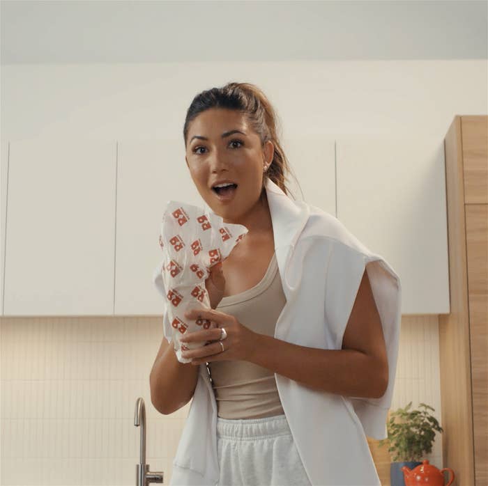 Woman in kitchen holding a playing card, looks surprised, casually dressed with a vest